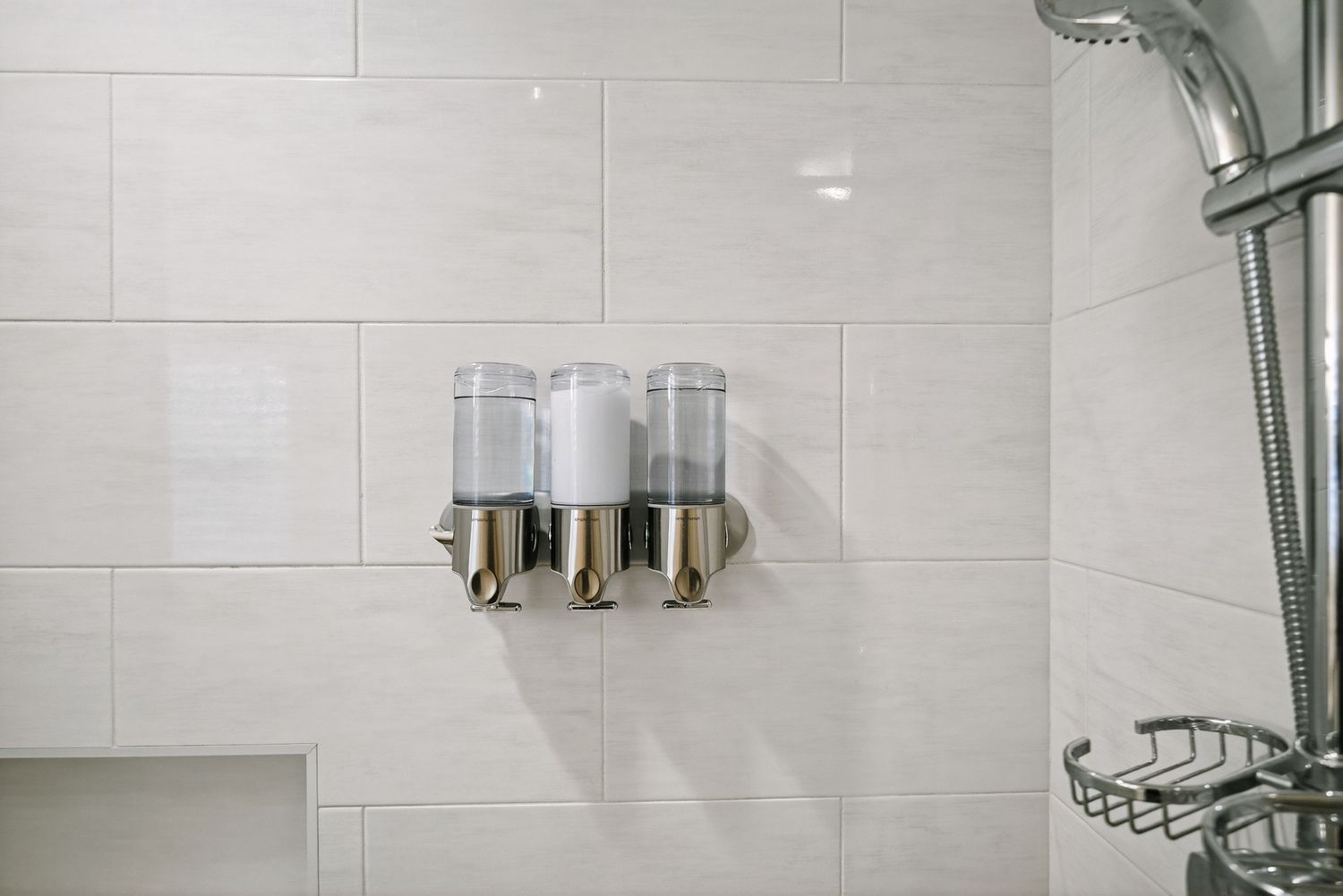 Modern dual wall-mounted soap dispensers in chrome finish installed on white tiled bathroom wall next to shower head.