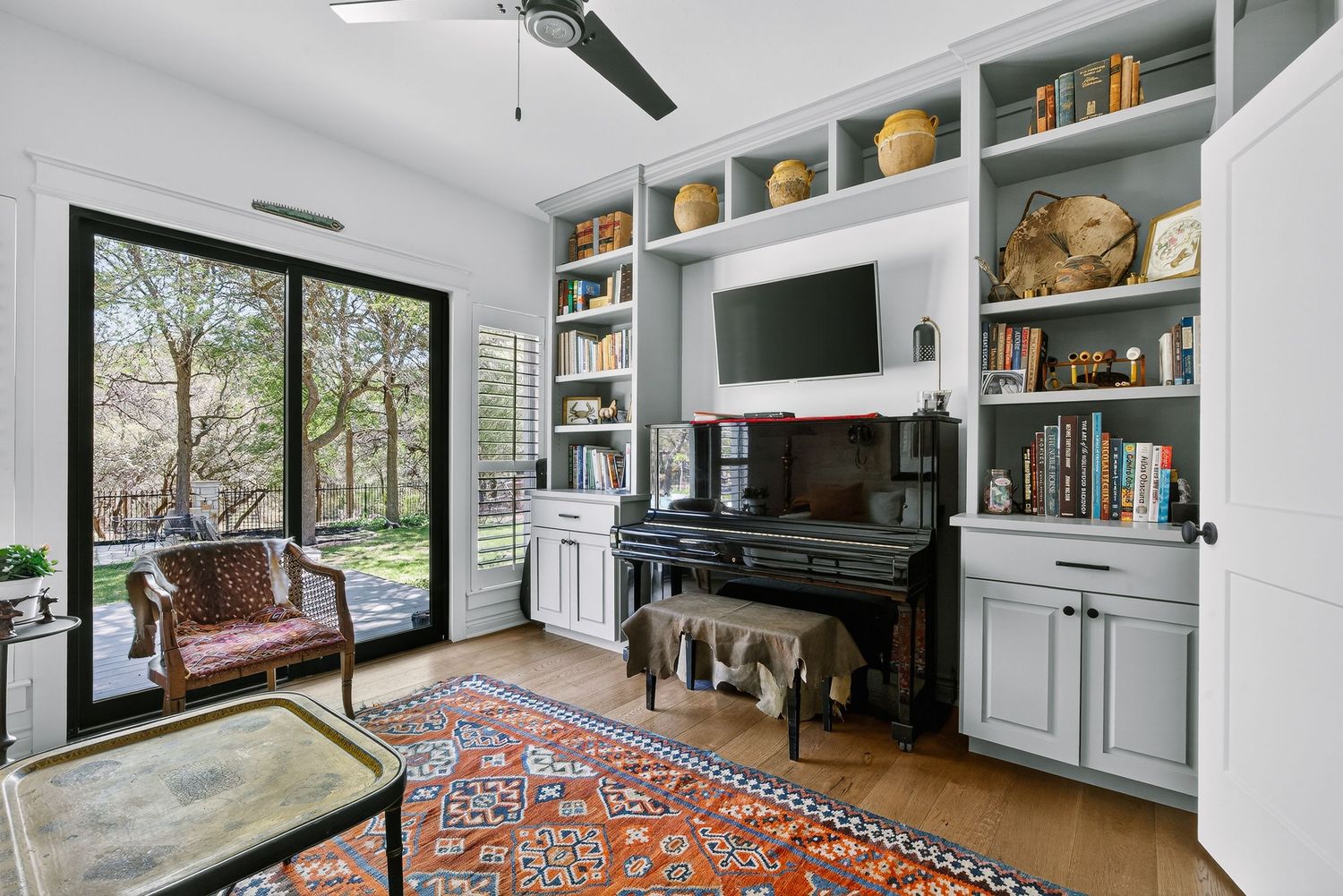 Cozy home office with built-in gray shelves, upright piano, wall-mounted TV, and sliding glass doors to a garden.