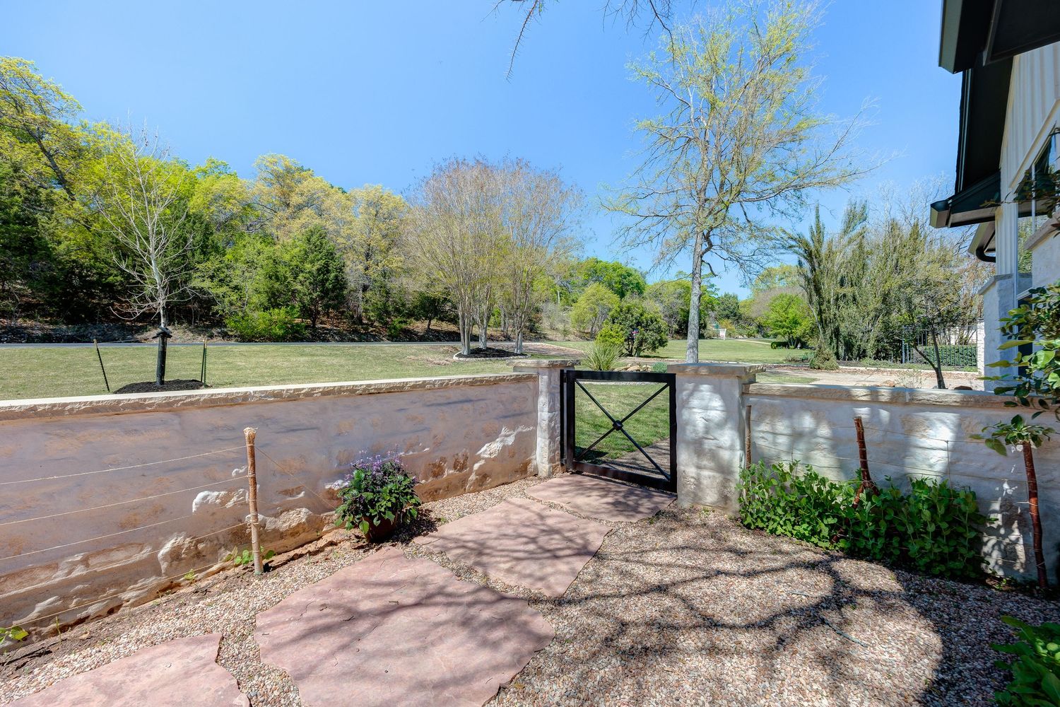 Rustic garden gate with white fence leads to a lush green yard with mature trees on a sunny day.