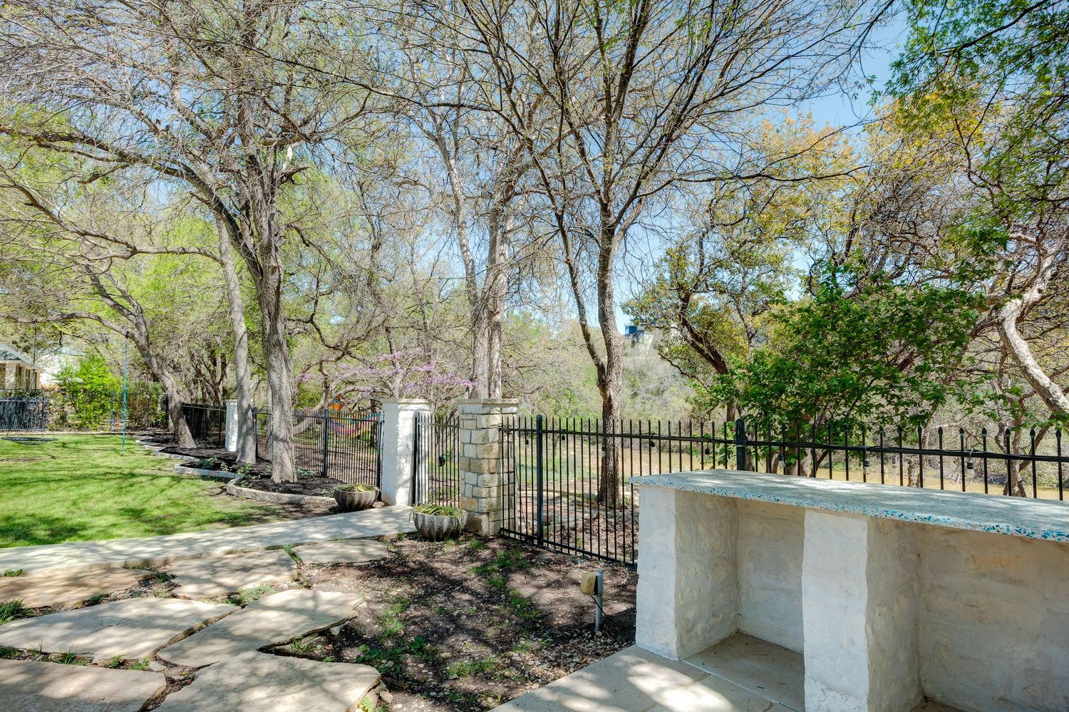 Stone patio with iron fence surrounded by mature trees and lush greenery in a serene outdoor garden setting.