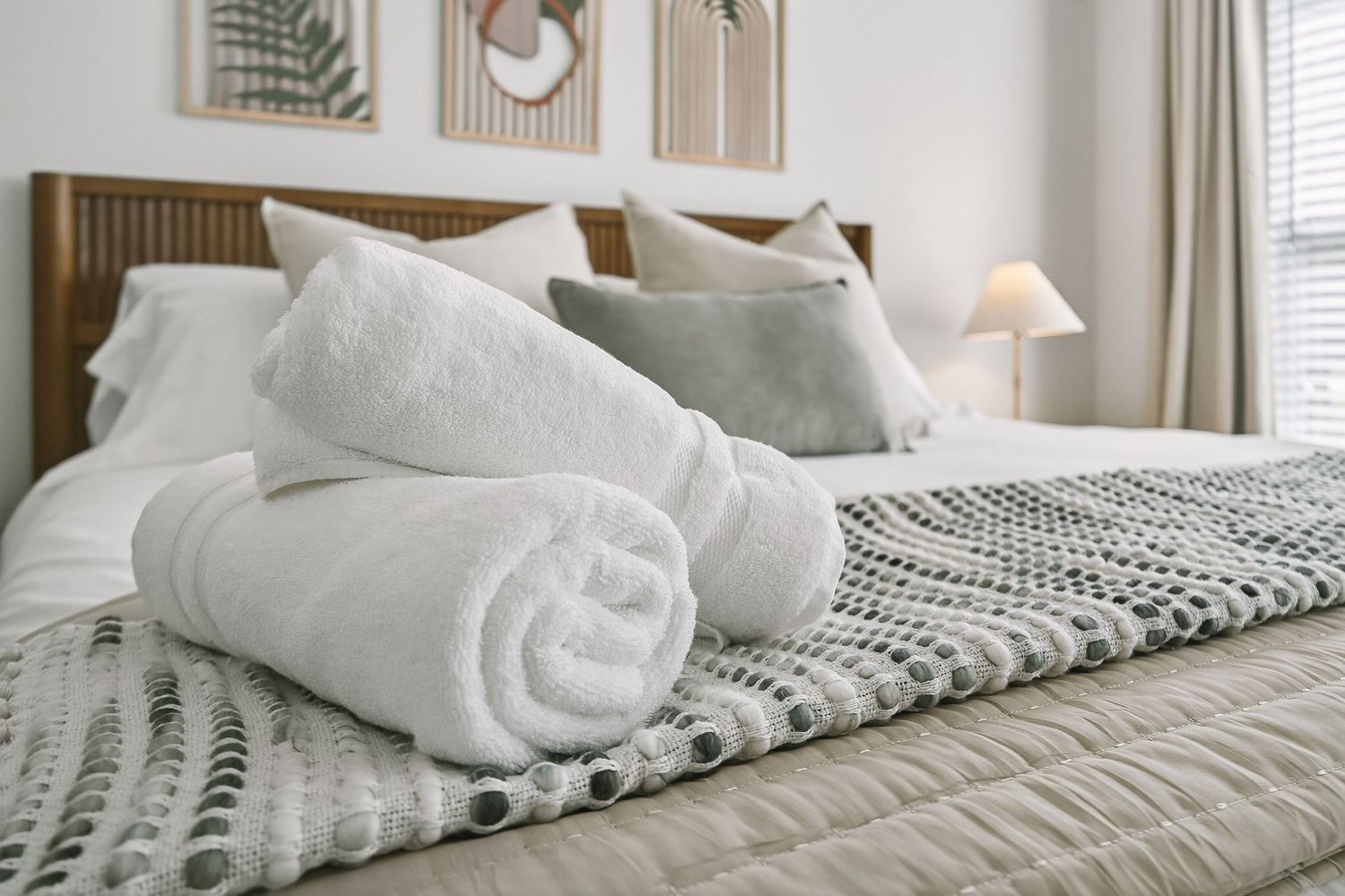 Cozy hotel bed with rolled white towels and knit throw blanket on neutral bedding with decorative wall art.