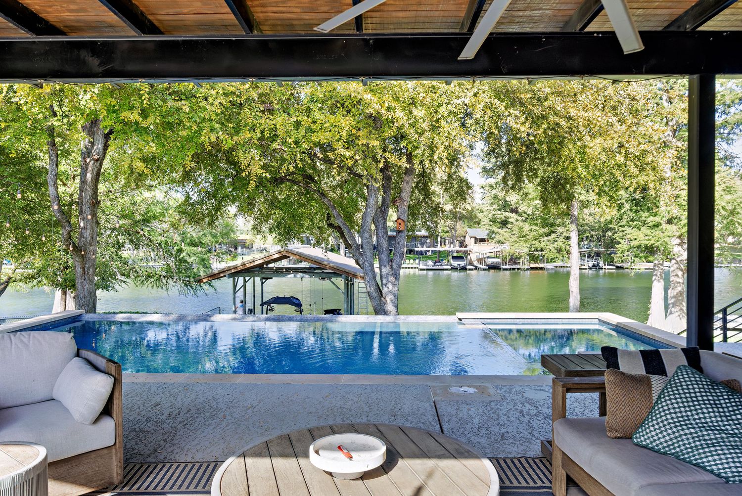 Covered lakeside deck with comfortable seating, an infinity pool, and tranquil water views through mature trees.