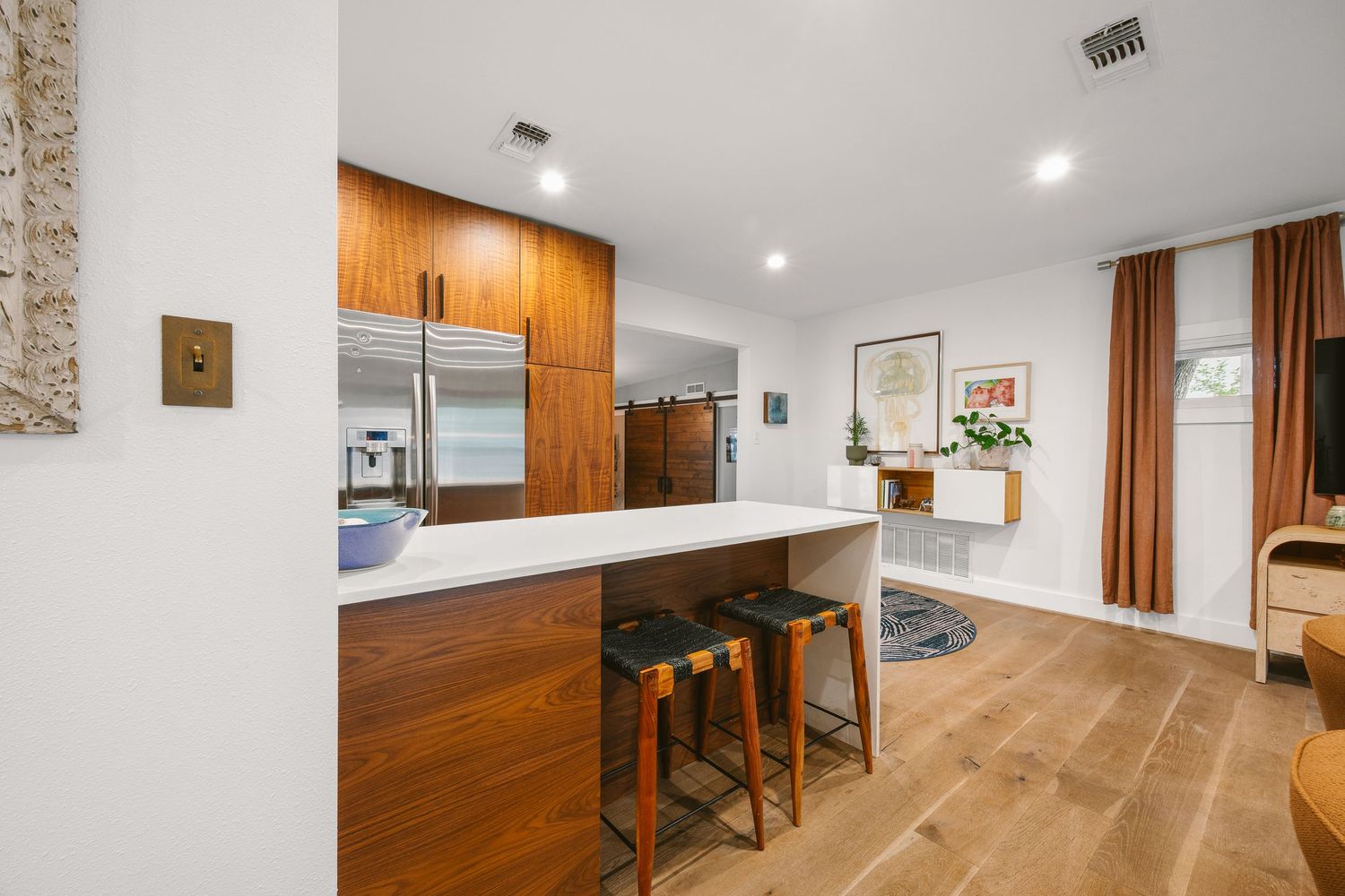 Modern kitchen with warm wood cabinetry, white countertop bar seating, stainless steel fridge, and light hardwood floors.
