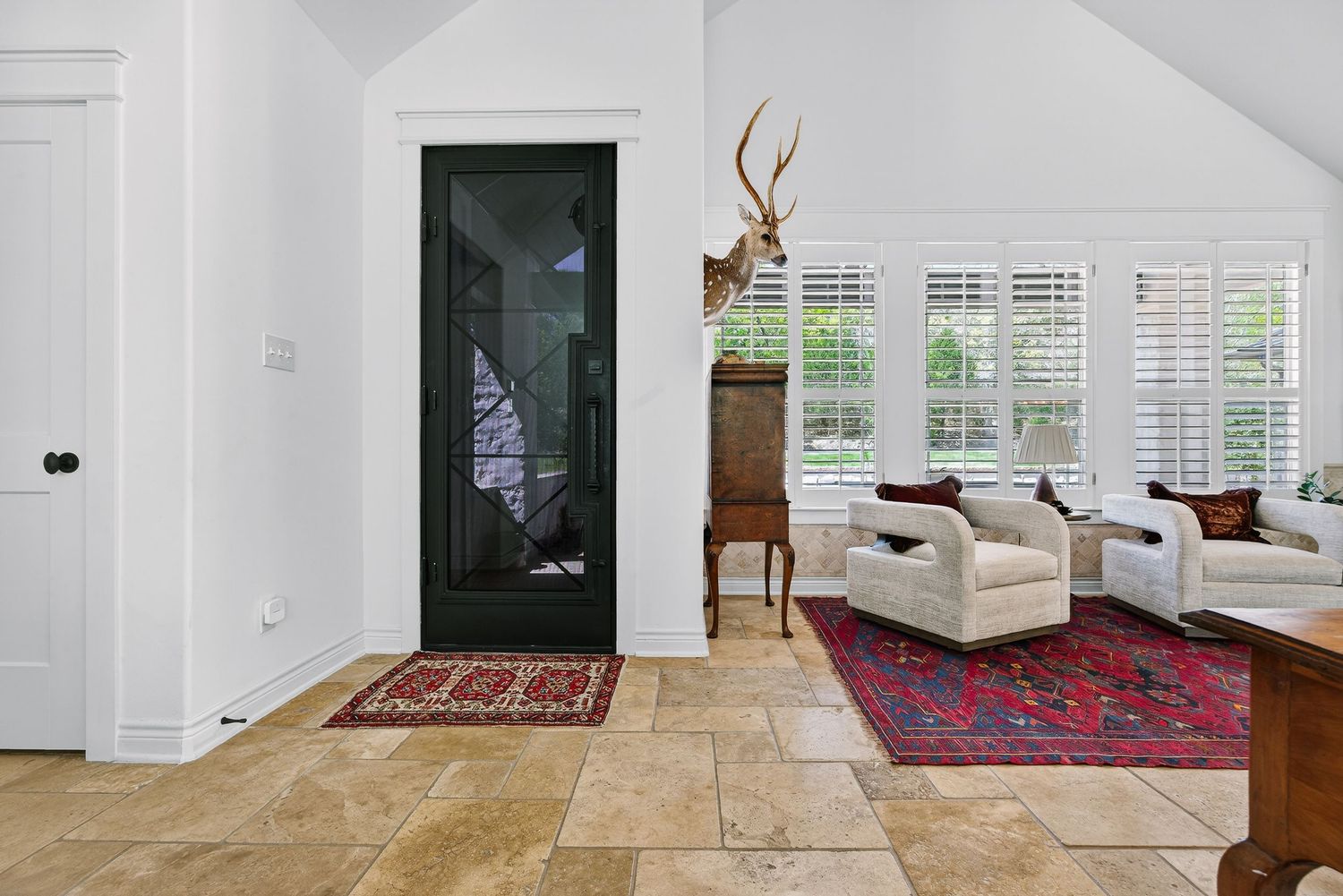 Bright living room with travertine floors, red Persian rug, white sofas, plantation shutters, and mounted deer antlers.