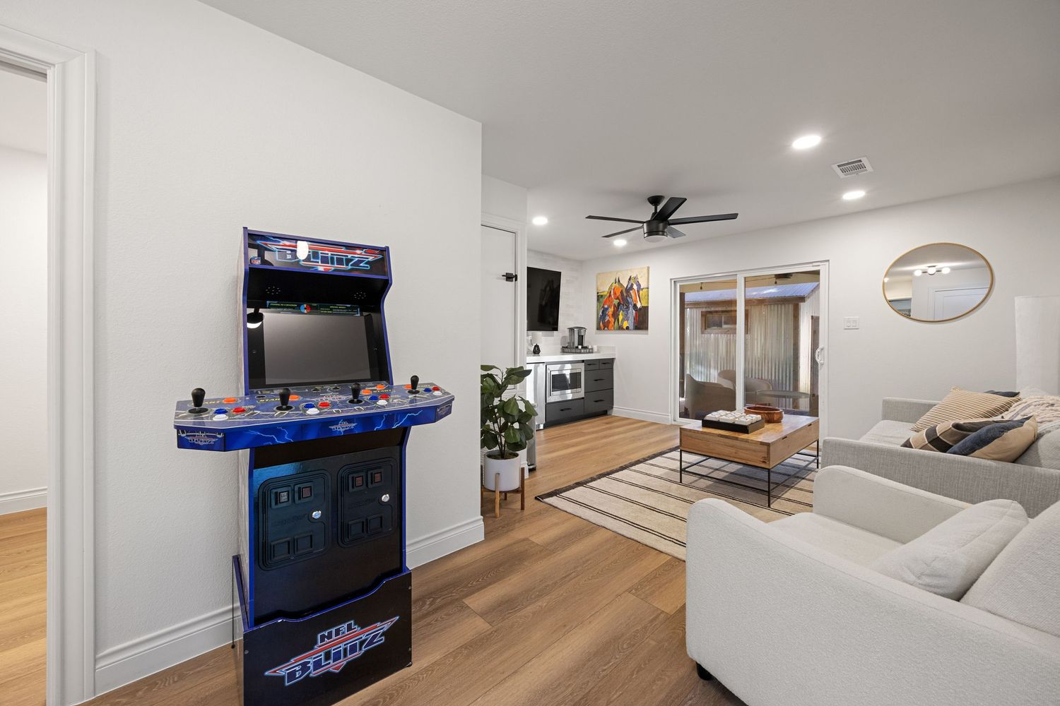 Bright living room with arcade game machine, hardwood floors, ceiling fan, and open layout leading to kitchen.
