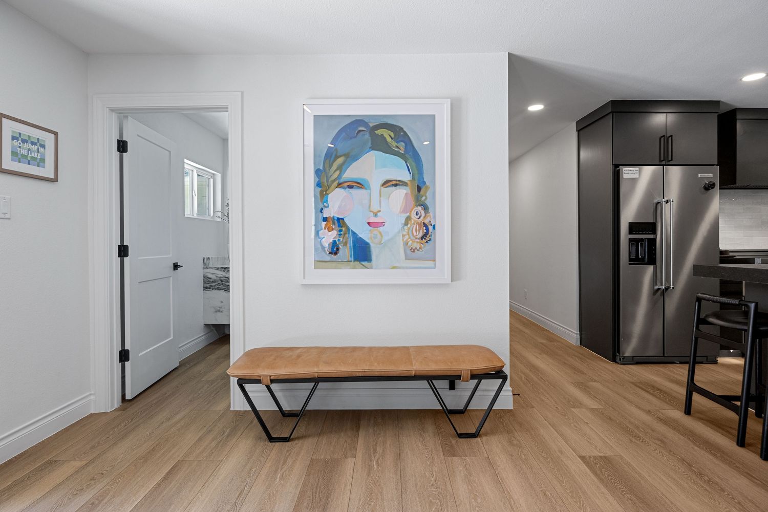 Modern entryway with wood bench, large portrait artwork, and stainless steel refrigerator on light hardwood floors.