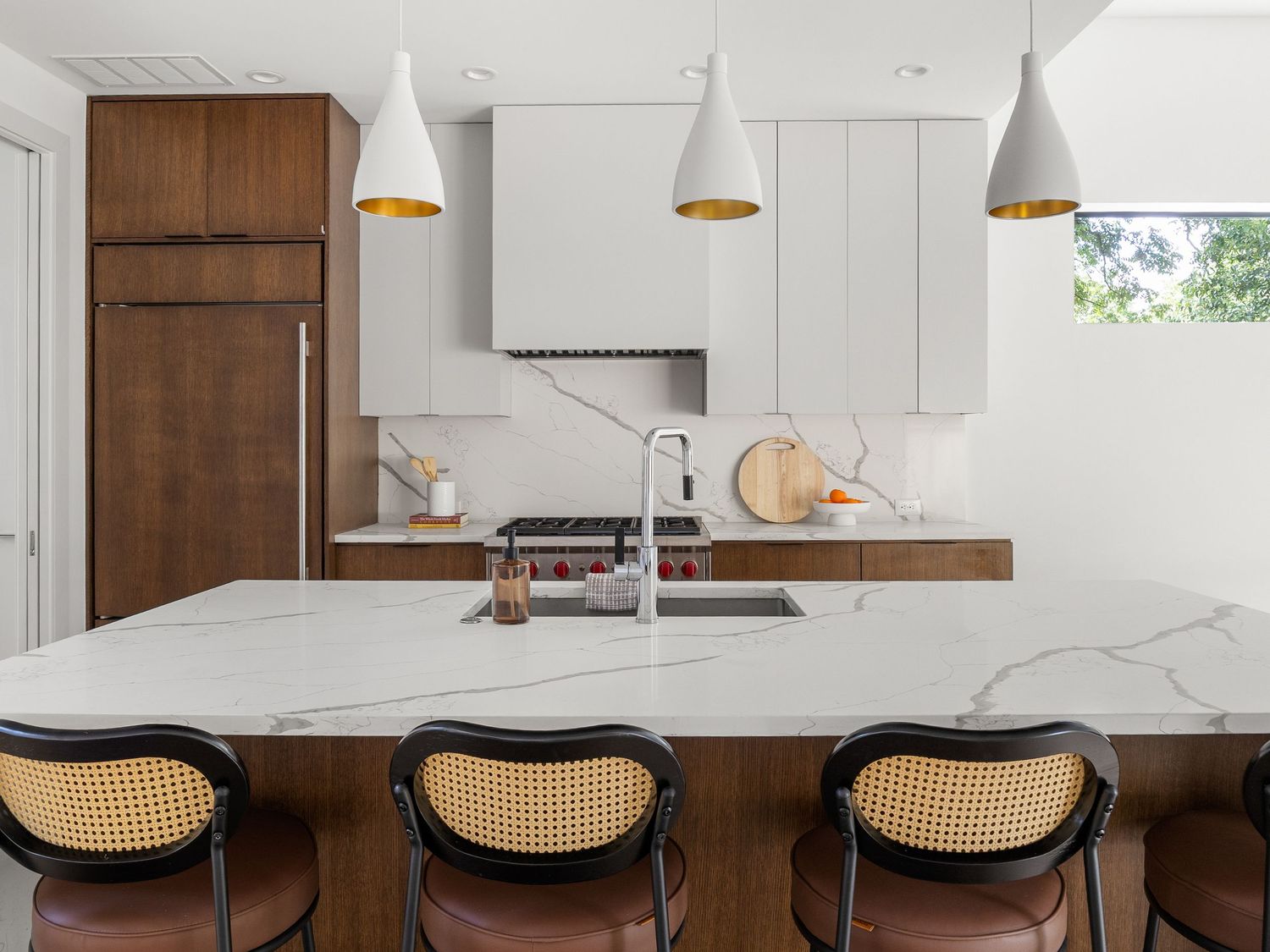 Modern kitchen with marble island, white pendant lights, walnut cabinets, and rattan bar stools in a bright open space.