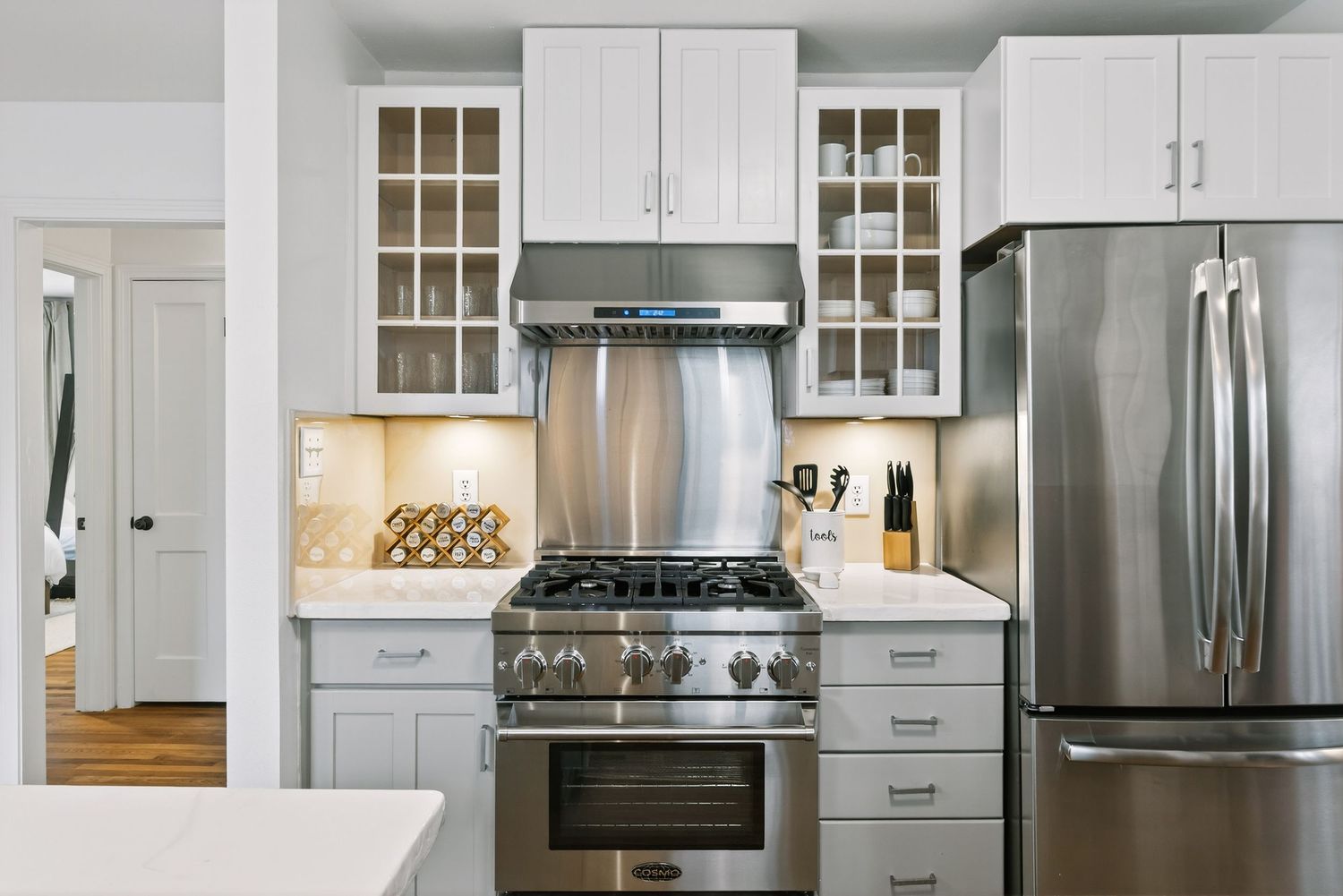 Modern white kitchen featuring stainless steel appliances, glass-front cabinets, and under-cabinet lighting.