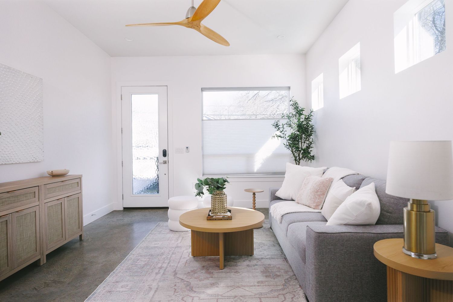 A bright minimalist living room with white walls, wooden furniture, and small windows letting in natural light.