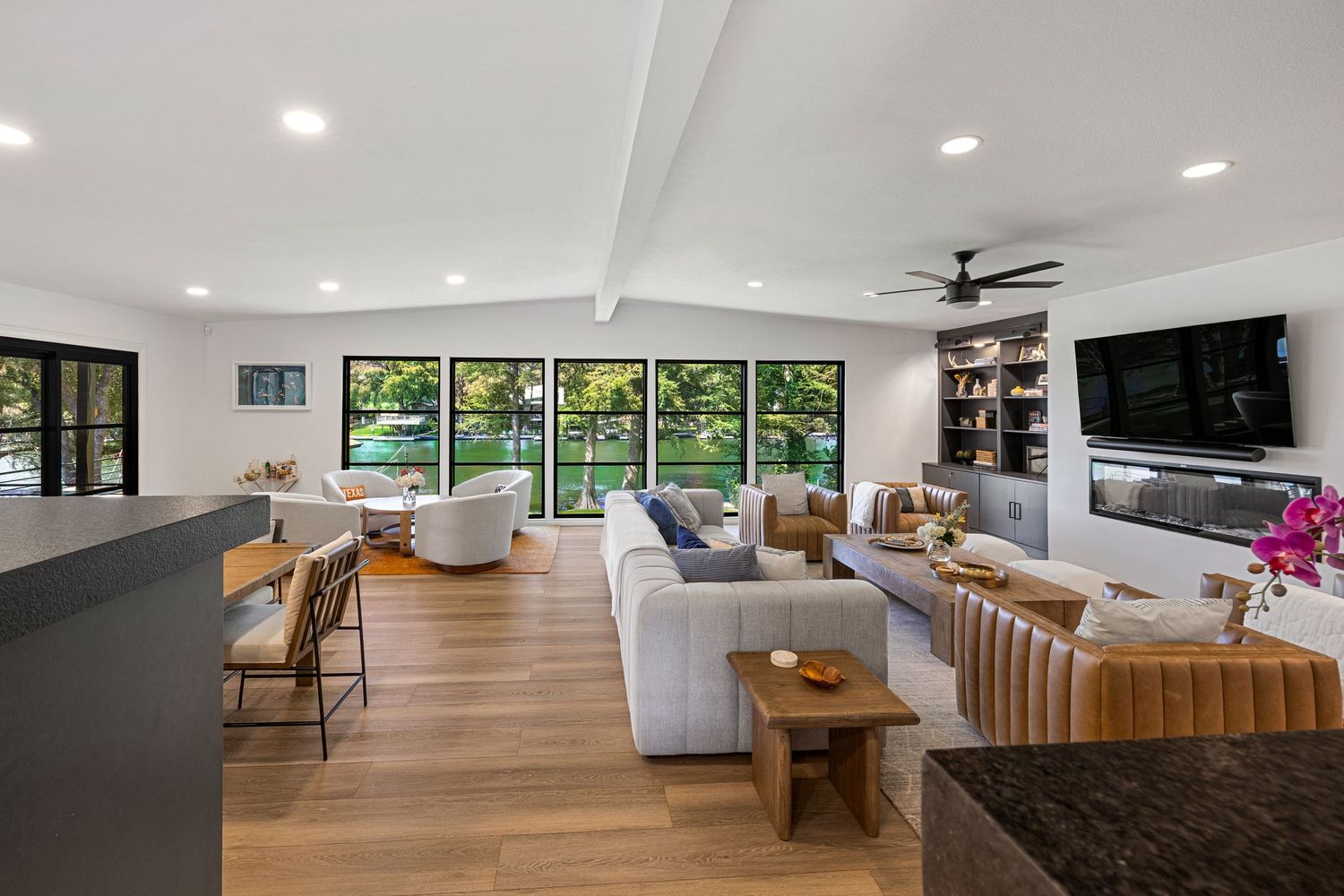 Open-concept living room with vaulted ceilings, built-in shelving, wall-mounted TV, and expansive windows with lush garden views.