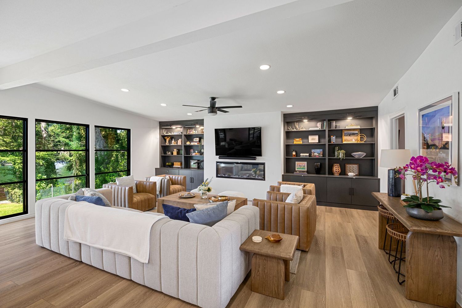 Spacious living room featuring built-in dark wood shelving, ceiling fan, fireplace, and light hardwood floors with neutral furnishings.