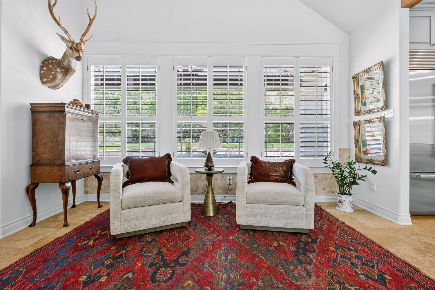 Elegant sitting room with vaulted ceiling, two white armchairs, red Persian rug, plantation shutters, and mounted deer head decor.