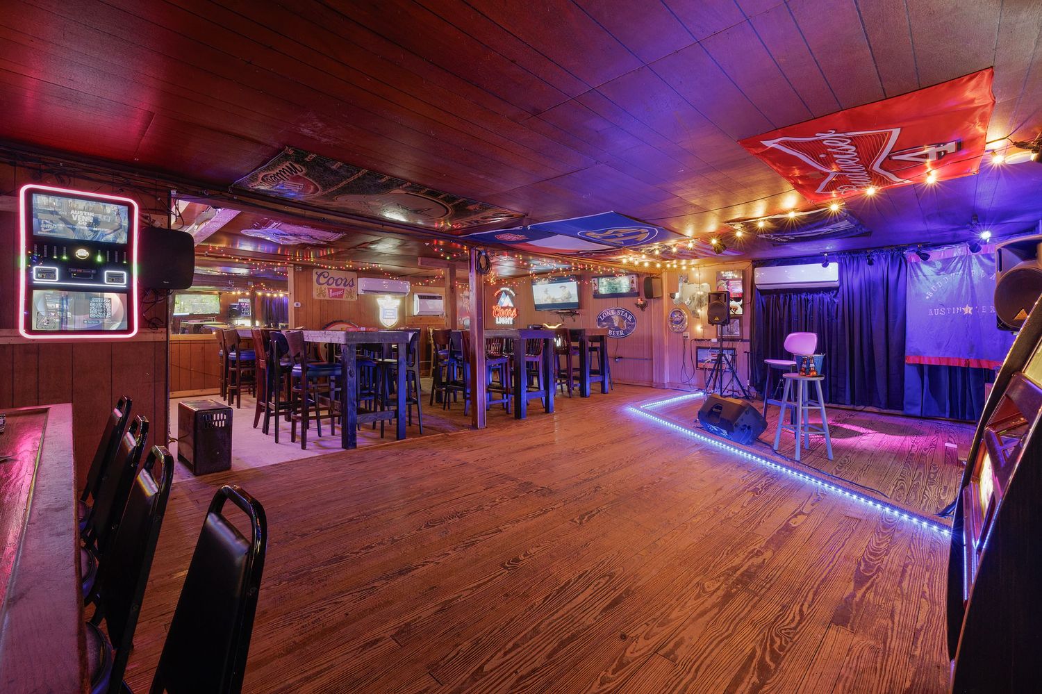 Spacious bar interior with hardwood floors, neon lighting, arcade games, high-top tables, and a stage area with purple LED accents.