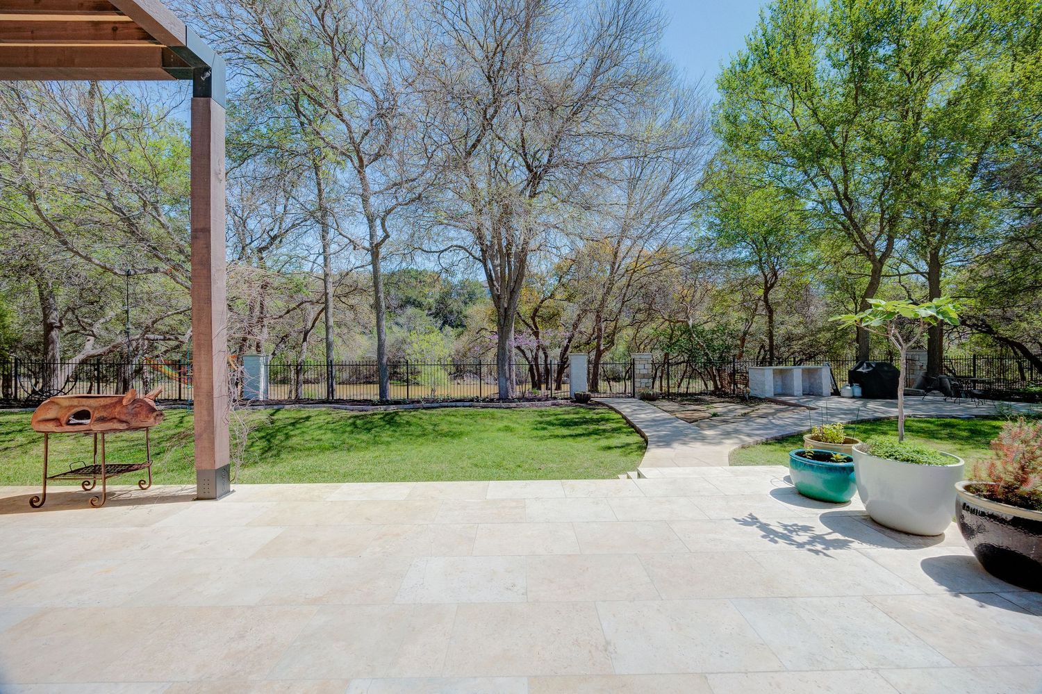 Spacious patio overlooking a lush green backyard with mature trees and a fenced garden area on a sunny day.