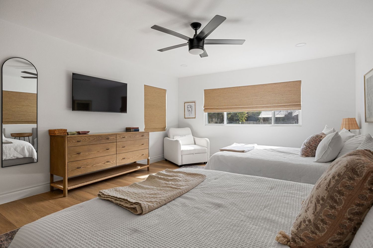 Spacious bedroom with two beds, ceiling fan, wall-mounted TV, wooden dresser, and natural light from bamboo shade windows.