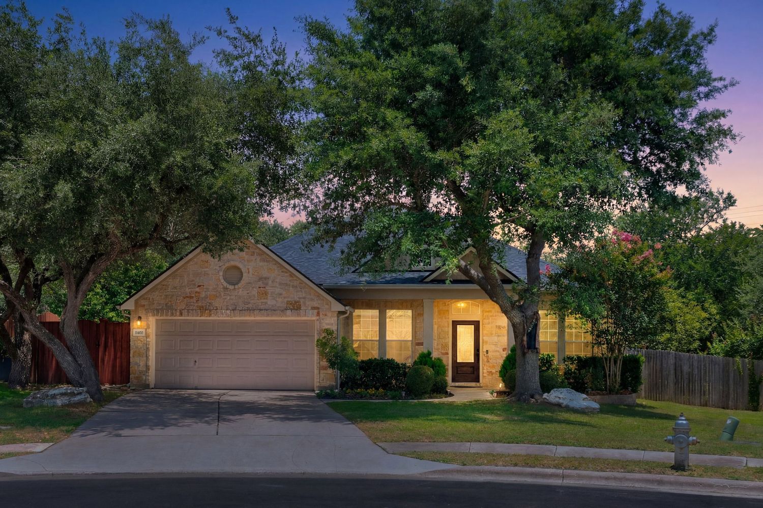 Single-story brick home with two-car garage surrounded by mature trees at twilight in a suburban neighborhood.