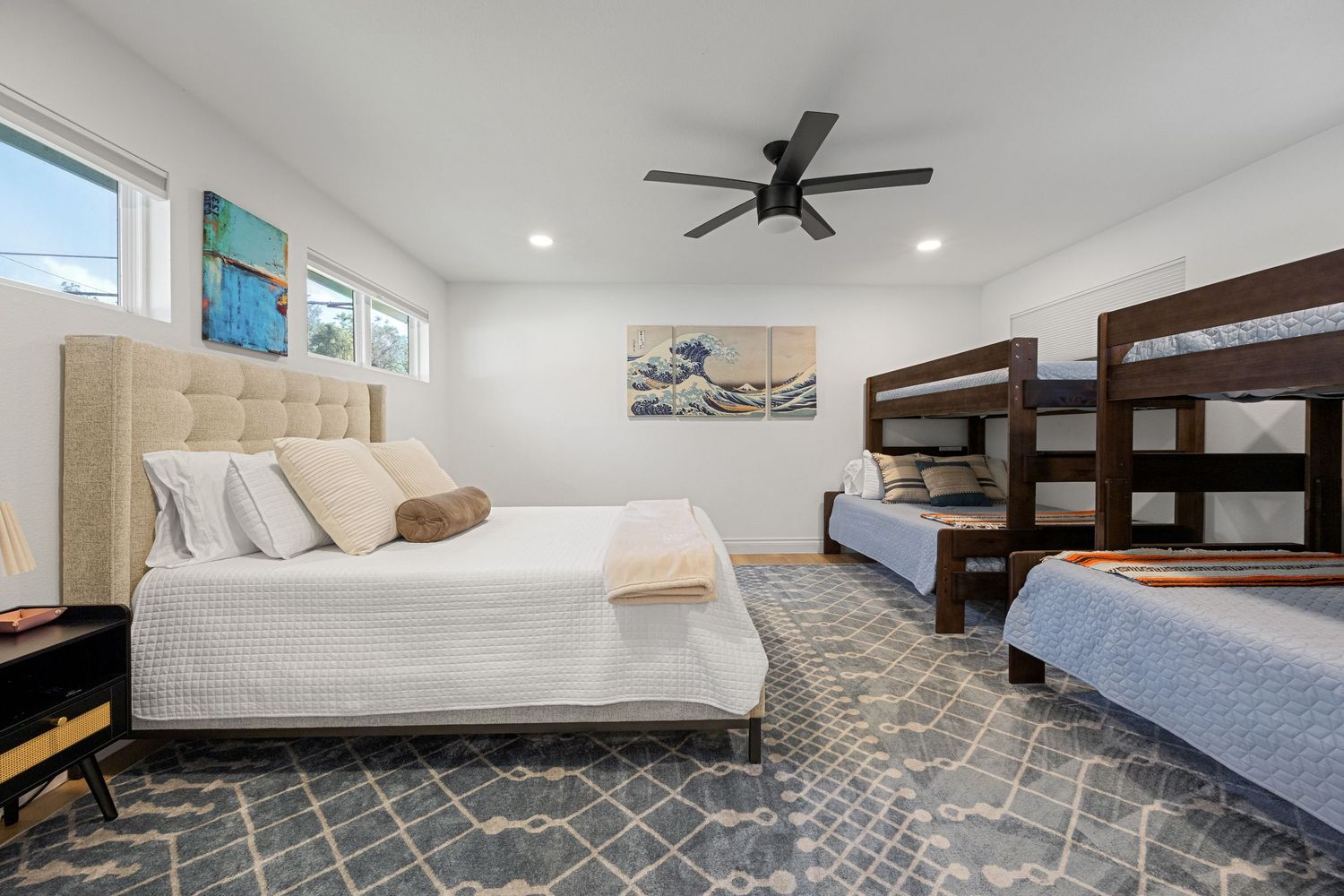 Spacious bedroom with a queen bed, wooden bunk bed, gray patterned area rug, ceiling fan, and coastal wall art on white walls.