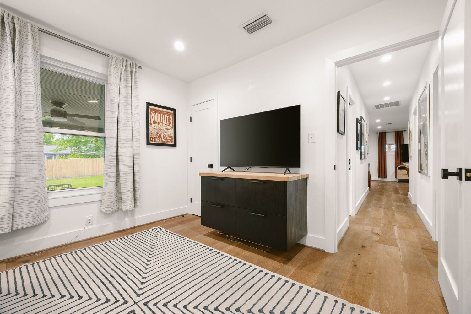 Modern living space with large TV on black dresser, hardwood floors, striped rug, and bright window with sheer curtains.