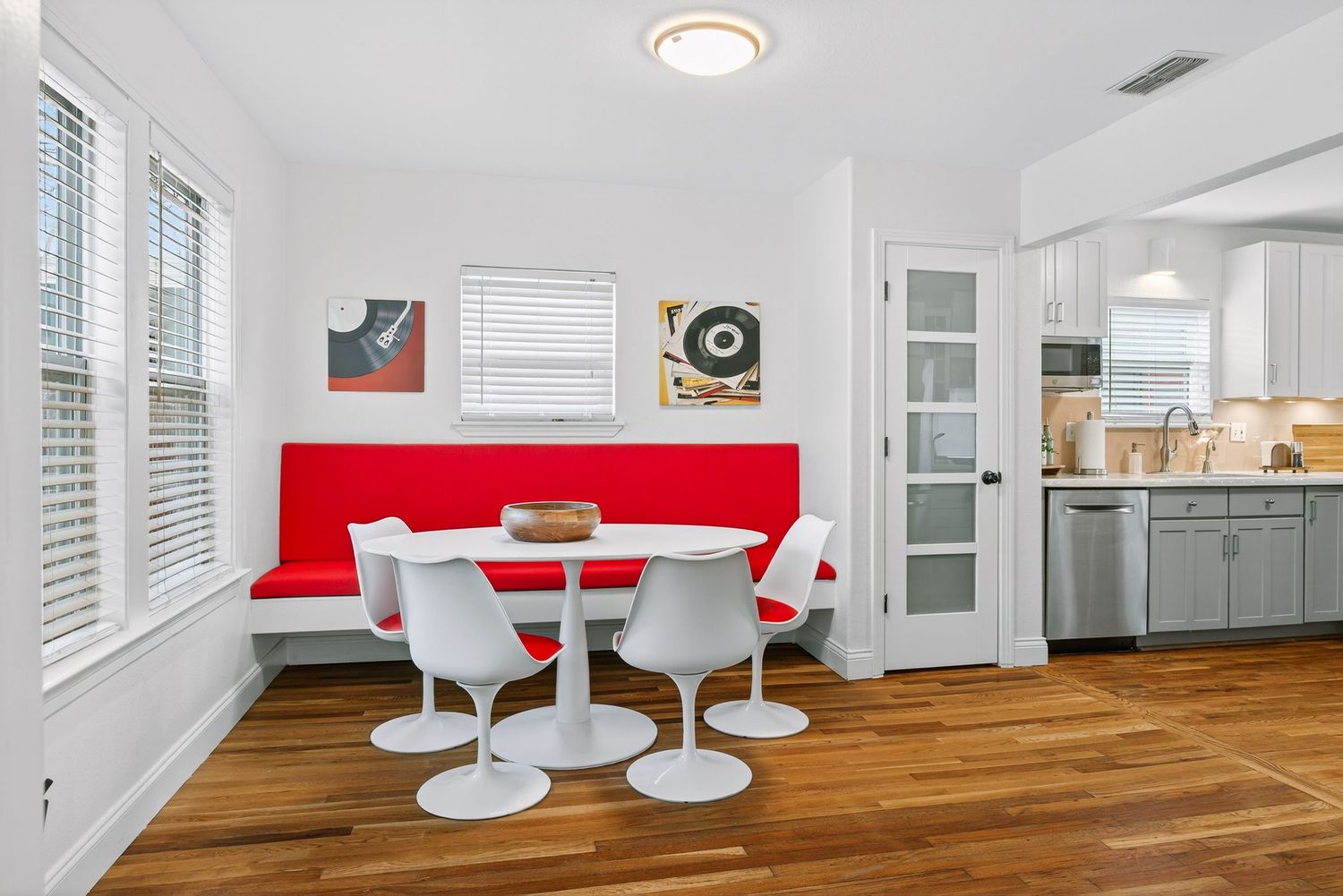Modern dining nook features a bright red banquette, white tulip table and chairs, with hardwood floors and kitchen view.