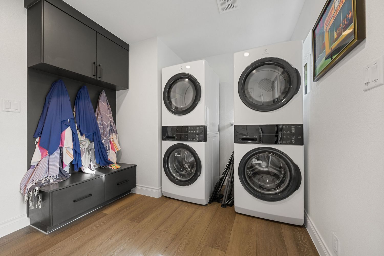 Modern laundry room with two stacked washer-dryer units, dark built-in storage bench, and hardwood flooring.