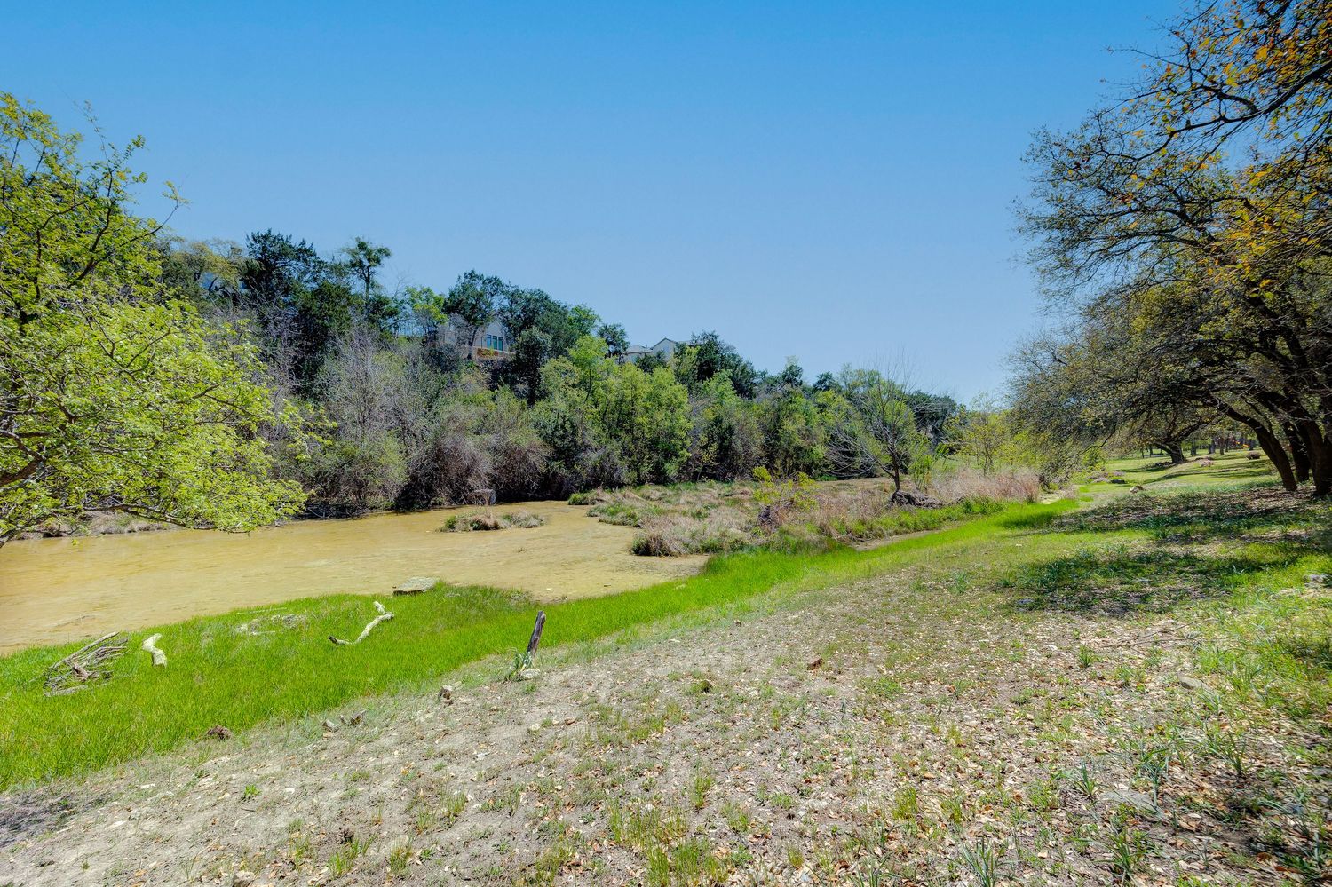 Scenic river landscape with lush green banks, muddy water, and dense tree line under a clear blue sky.