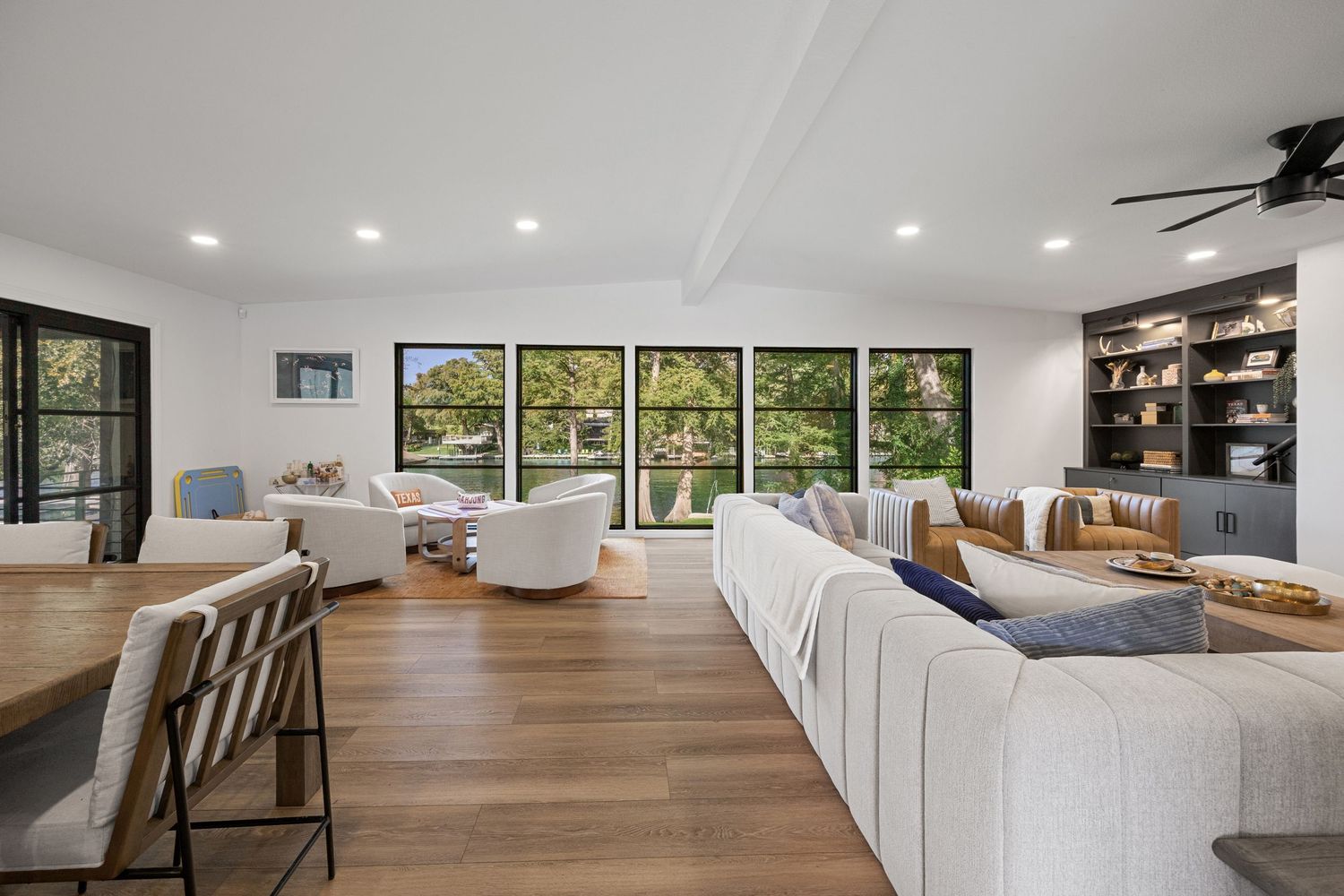 Bright open-concept living room with white sectional sofa, hardwood floors, large windows, ceiling fan, and built-in shelving.