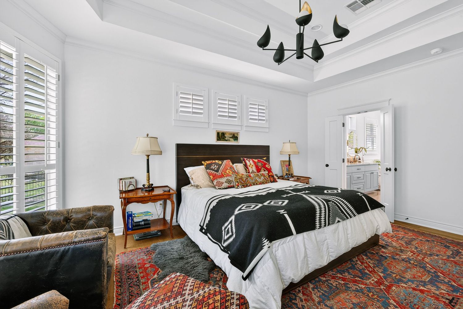 Elegant bedroom with tray ceiling, black chandelier, bold patterned bedding, plantation shutters, and vibrant red area rug.