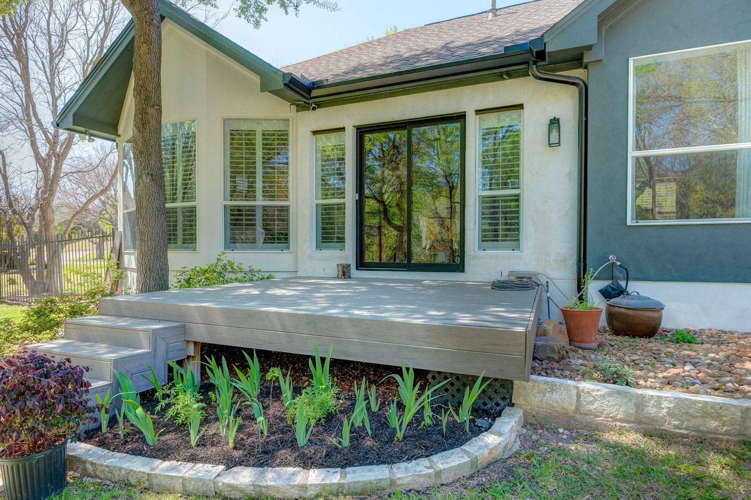 Modern home exterior with wood deck, large black sliding door, and curved garden bed with green plants.