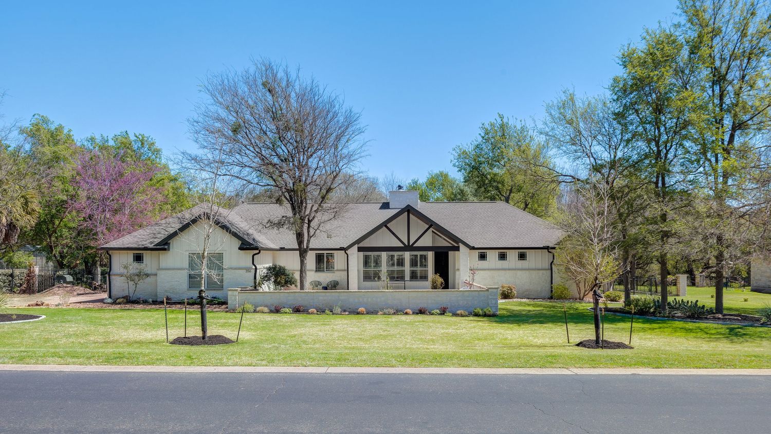 Single-story ranch-style home with white brick exterior, Tudor accents, lush green lawn, and mature trees on a sunny day.