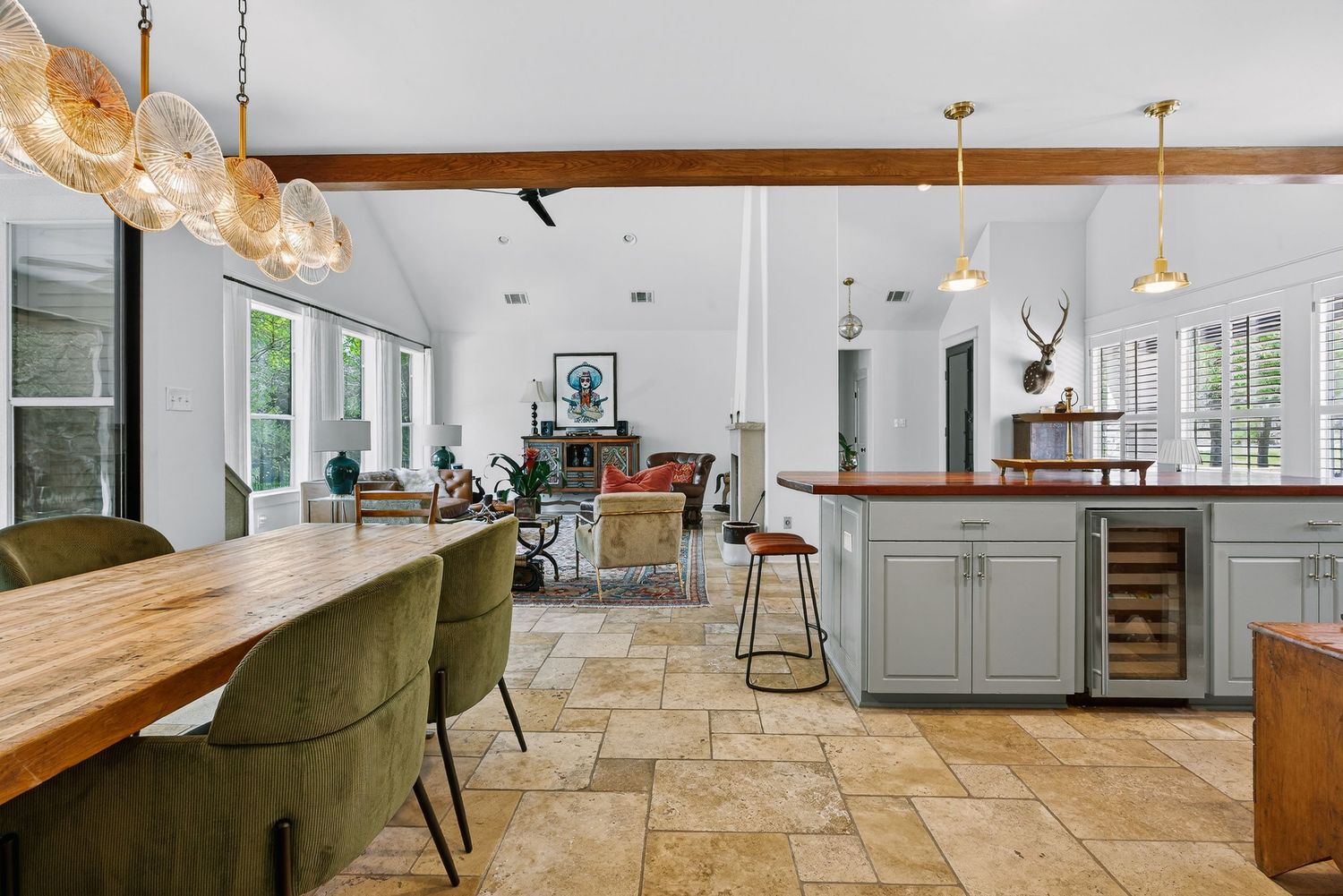 Open-concept kitchen and dining area with stone tile floors, green chairs, gray cabinets, wood countertops, and exposed ceiling beams.