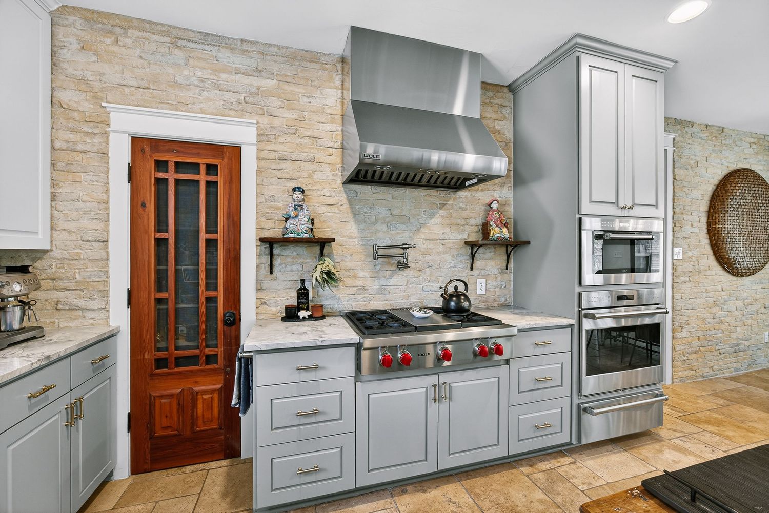 Modern kitchen with gray cabinets, stone backsplash, stainless steel range hood, and gas cooktop near a wooden door.