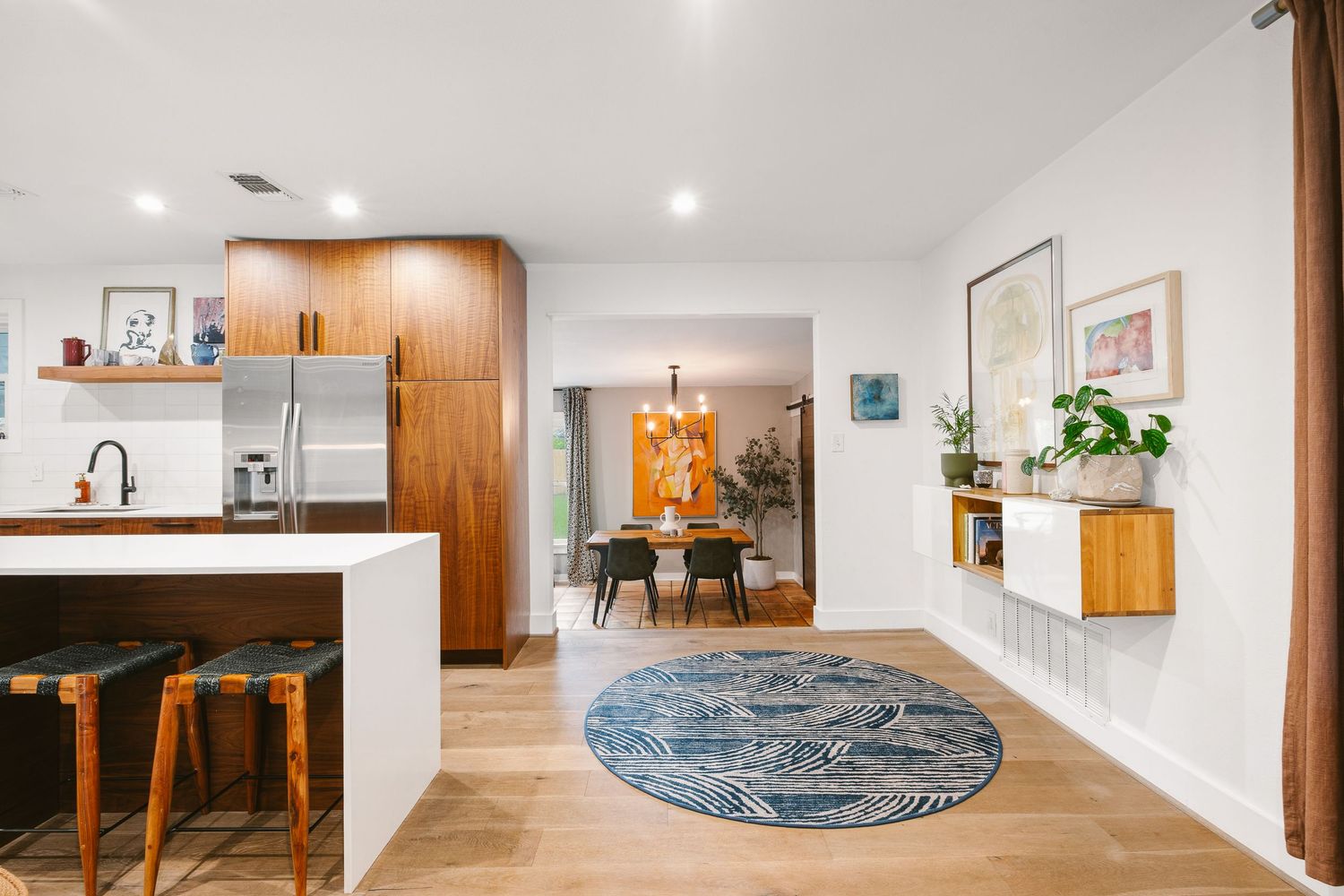 Modern open-plan kitchen and living area with wood cabinetry, round blue rug, bar stools, and dining room visible in background.