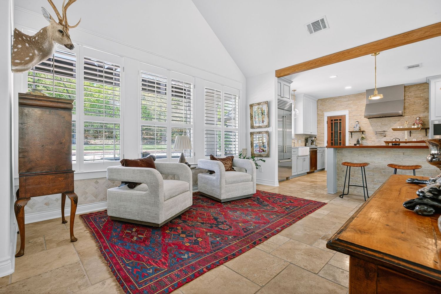 Bright living room with vaulted ceilings, white chairs, red Persian rug, and open kitchen with bar seating in background.