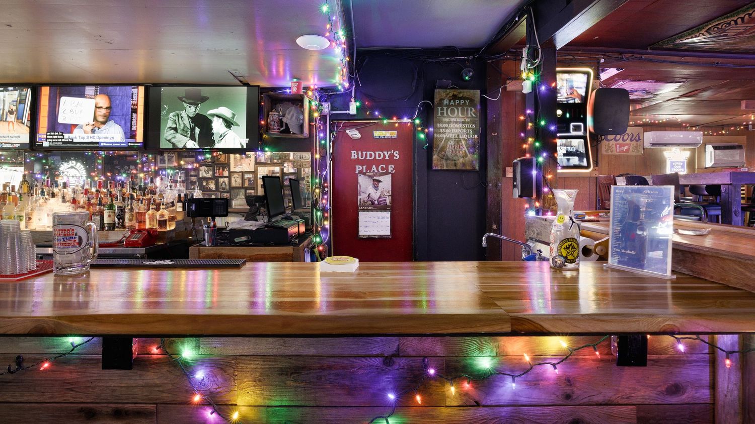 Cozy bar interior decorated with colorful string lights along the wooden counter, bottles displayed on shelves, and TVs mounted on the wall.