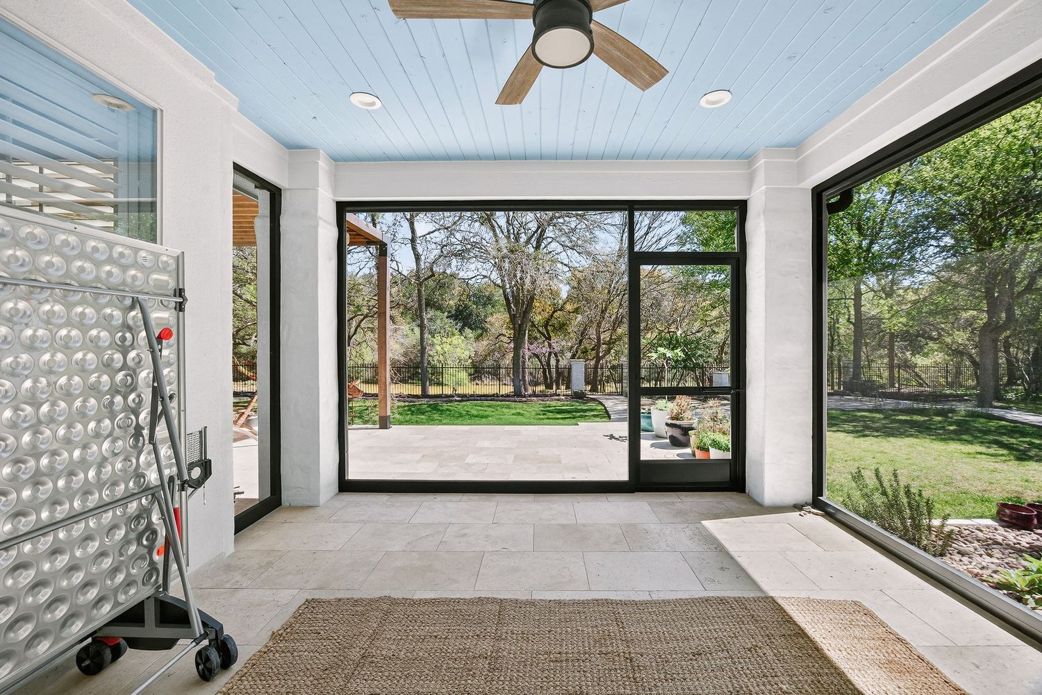 Bright sunroom with ceiling fan, large glass doors, and scenic backyard views of lush trees and green lawn.
