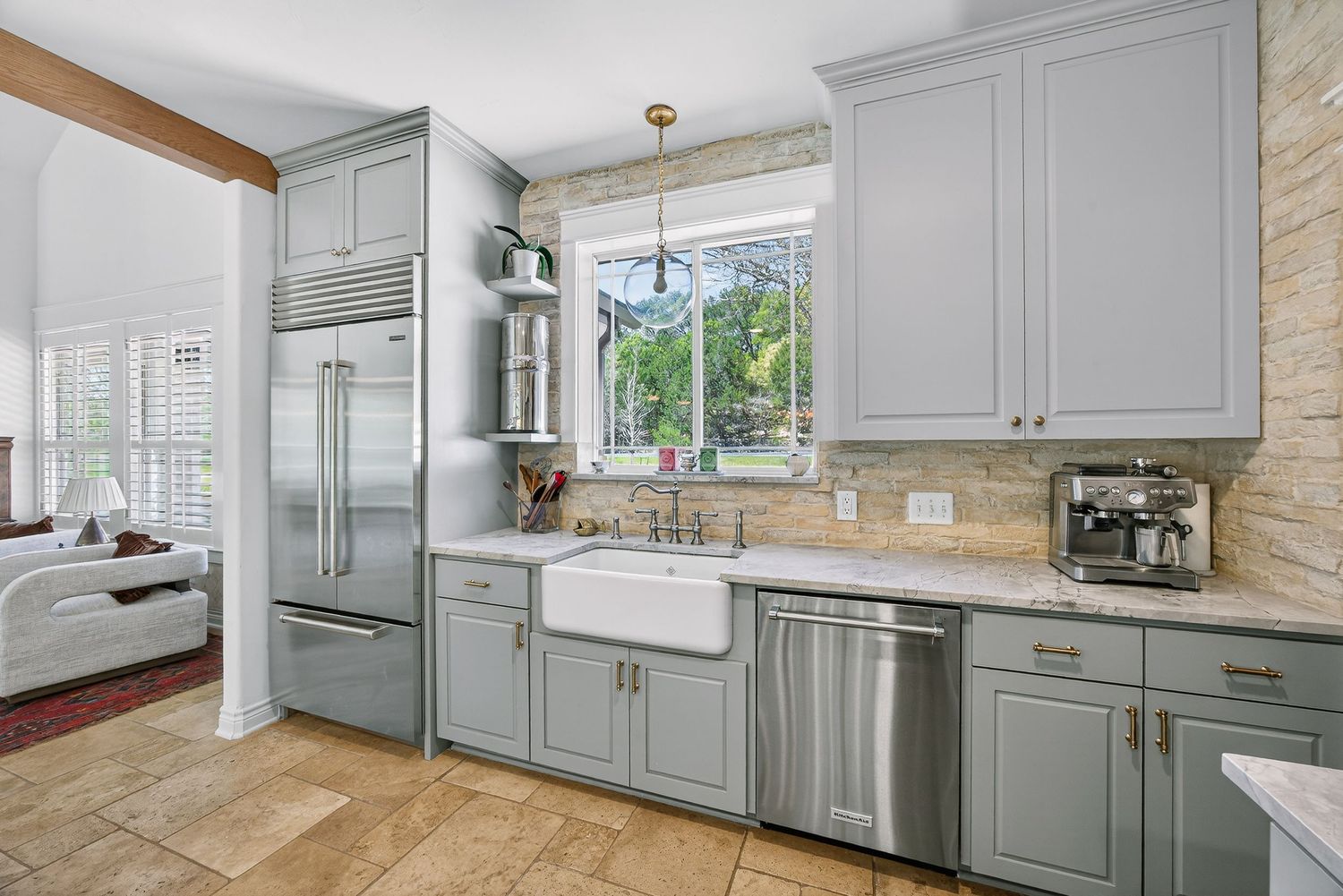 Gray kitchen cabinets with farmhouse sink, stainless appliances, stone backsplash, and pendant lighting over tile floors.