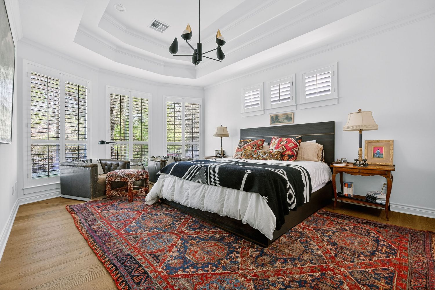 Elegant bedroom with tray ceiling, black chandelier, dark wood bed, vibrant Persian rug, and plantation shutters on large windows.