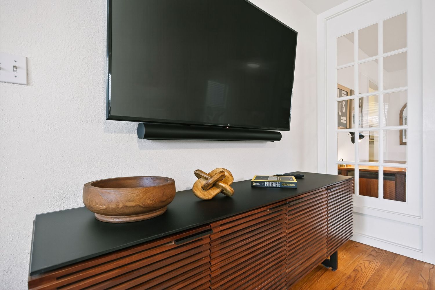 Modern living room setup with wall-mounted TV above wooden media console featuring slatted design and decorative bowl.