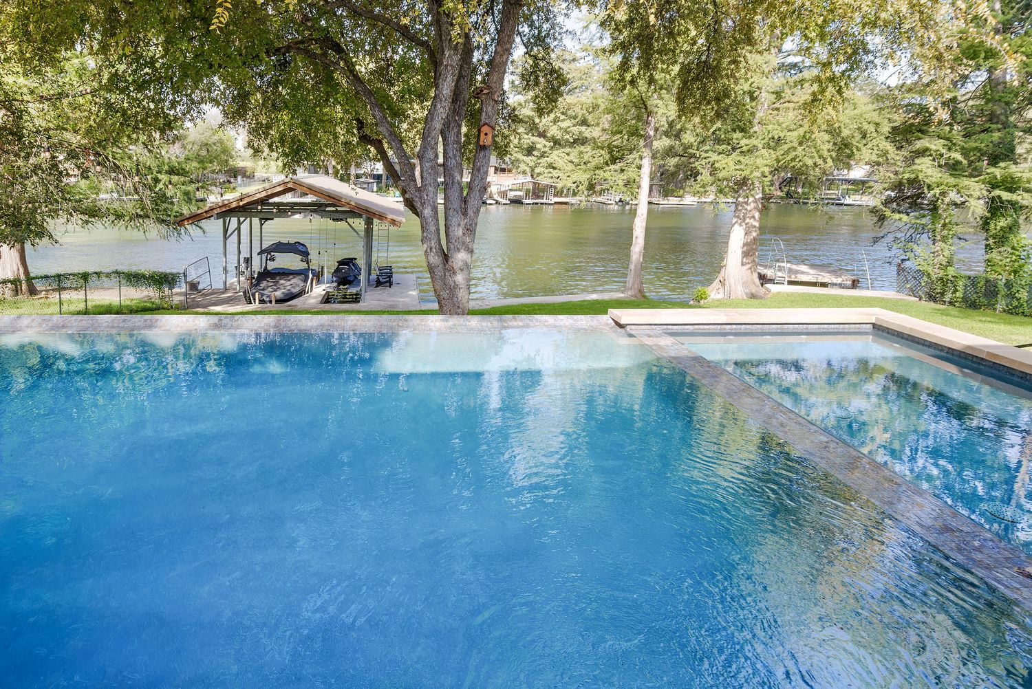 Stunning infinity pool with serene waterfront views, lush trees, and a covered boat dock in the background.
