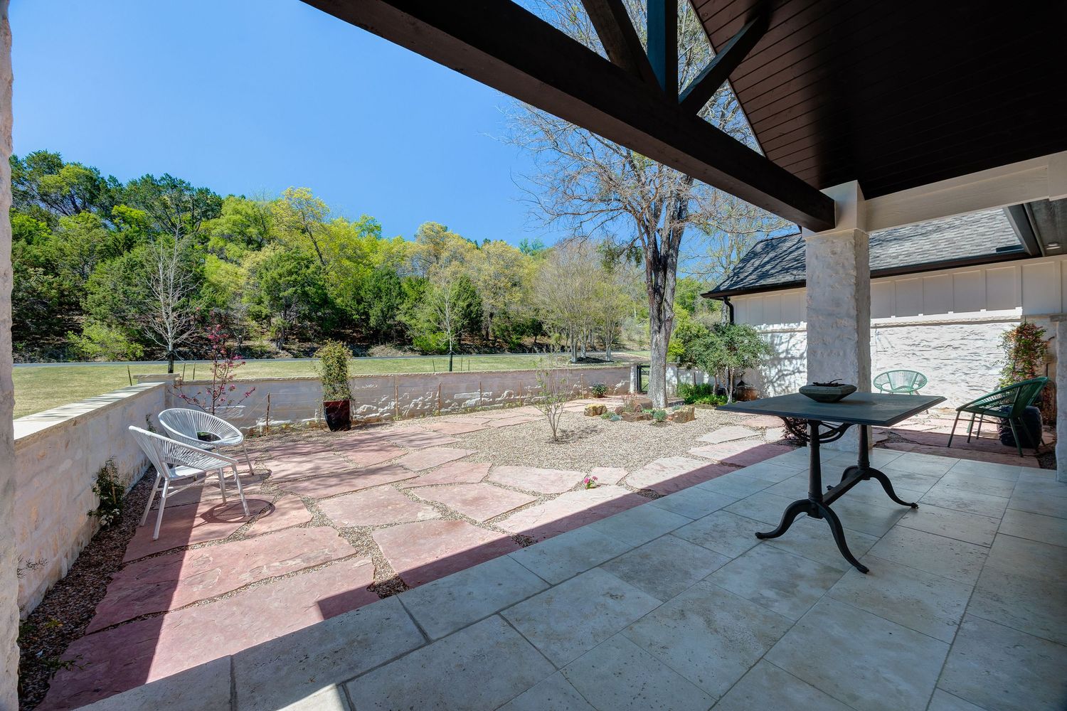 Covered patio with stone flooring overlooking a lush green backyard with trees and a decorative bistro table.