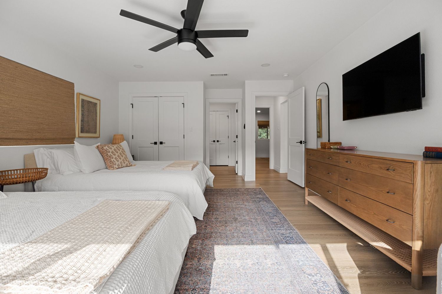 Elegant master bedroom with two queen beds, natural wood headboards, dresser, wall-mounted TV, and light hardwood floors.