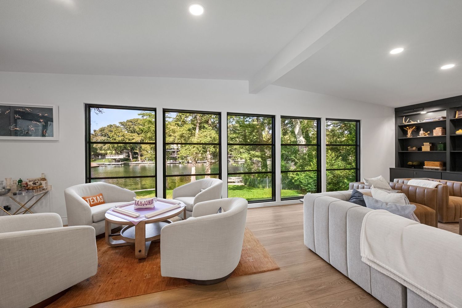 Modern living room with floor-to-ceiling black-framed windows overlooking a serene pond, featuring white sofas and hardwood floors.