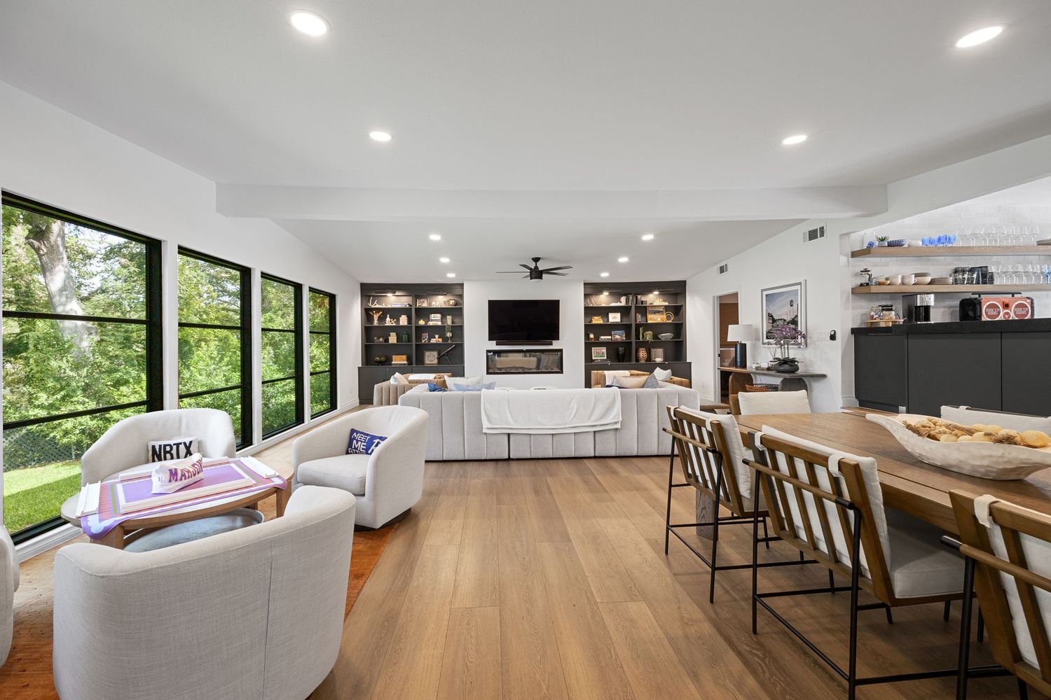 Bright open-concept living room with white sofas, built-in bookshelves, dining area, and large windows overlooking lush greenery.
