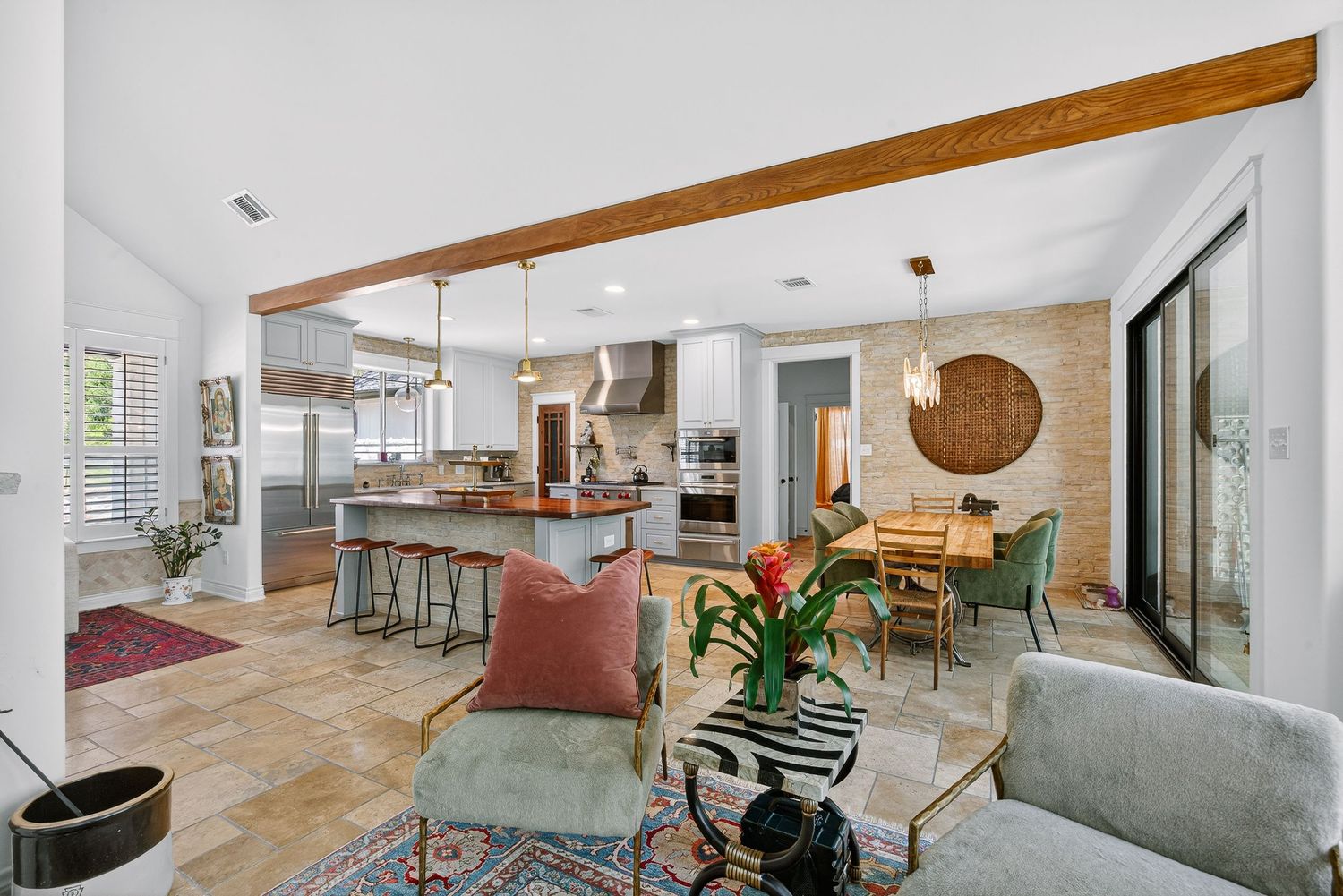 Open-concept living room and kitchen with exposed wood beams, colorful rug, island seating, and sliding glass doors to patio.