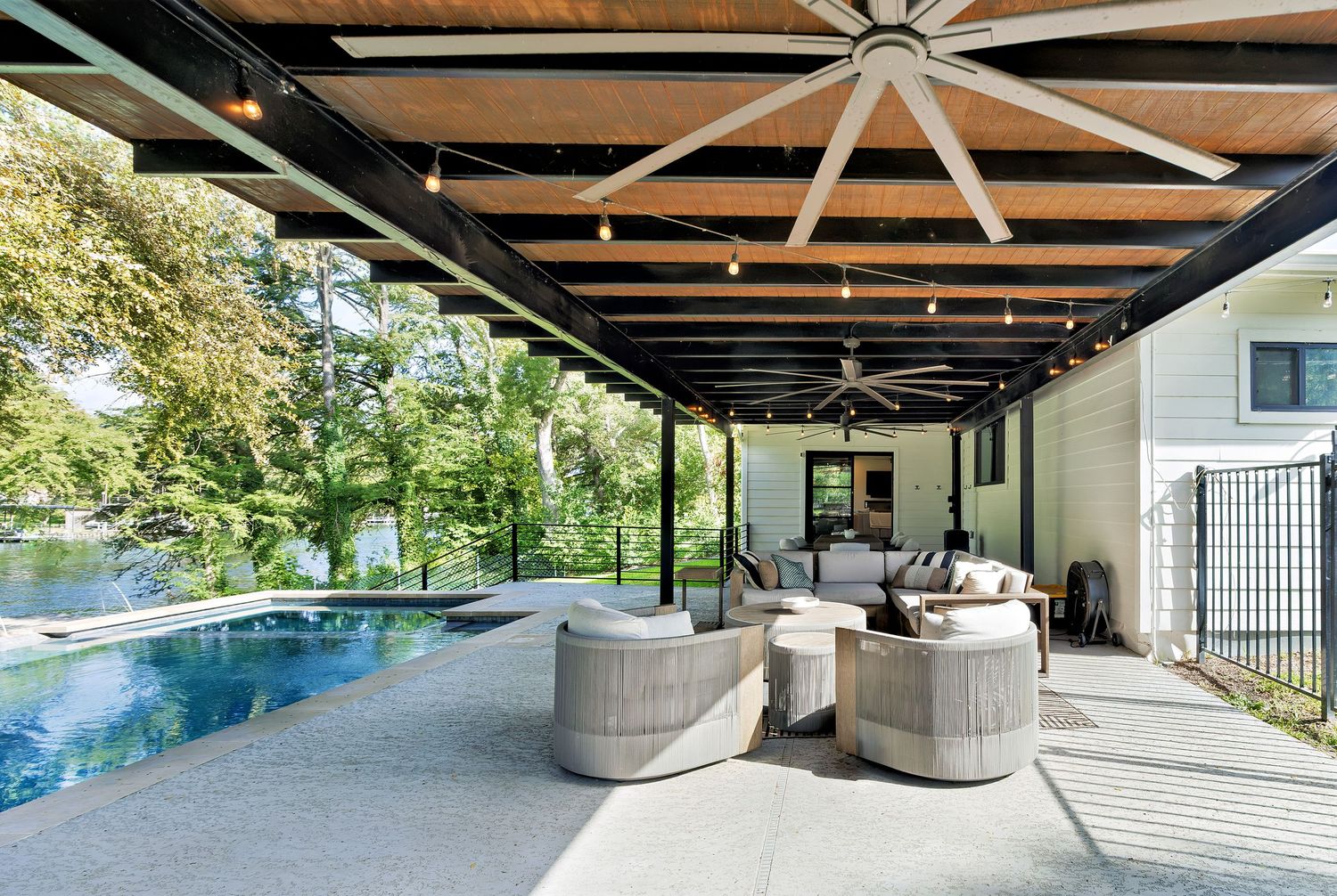 Covered outdoor patio with large ceiling fan, string lights, pool, lounge seating, and lush waterfront views.