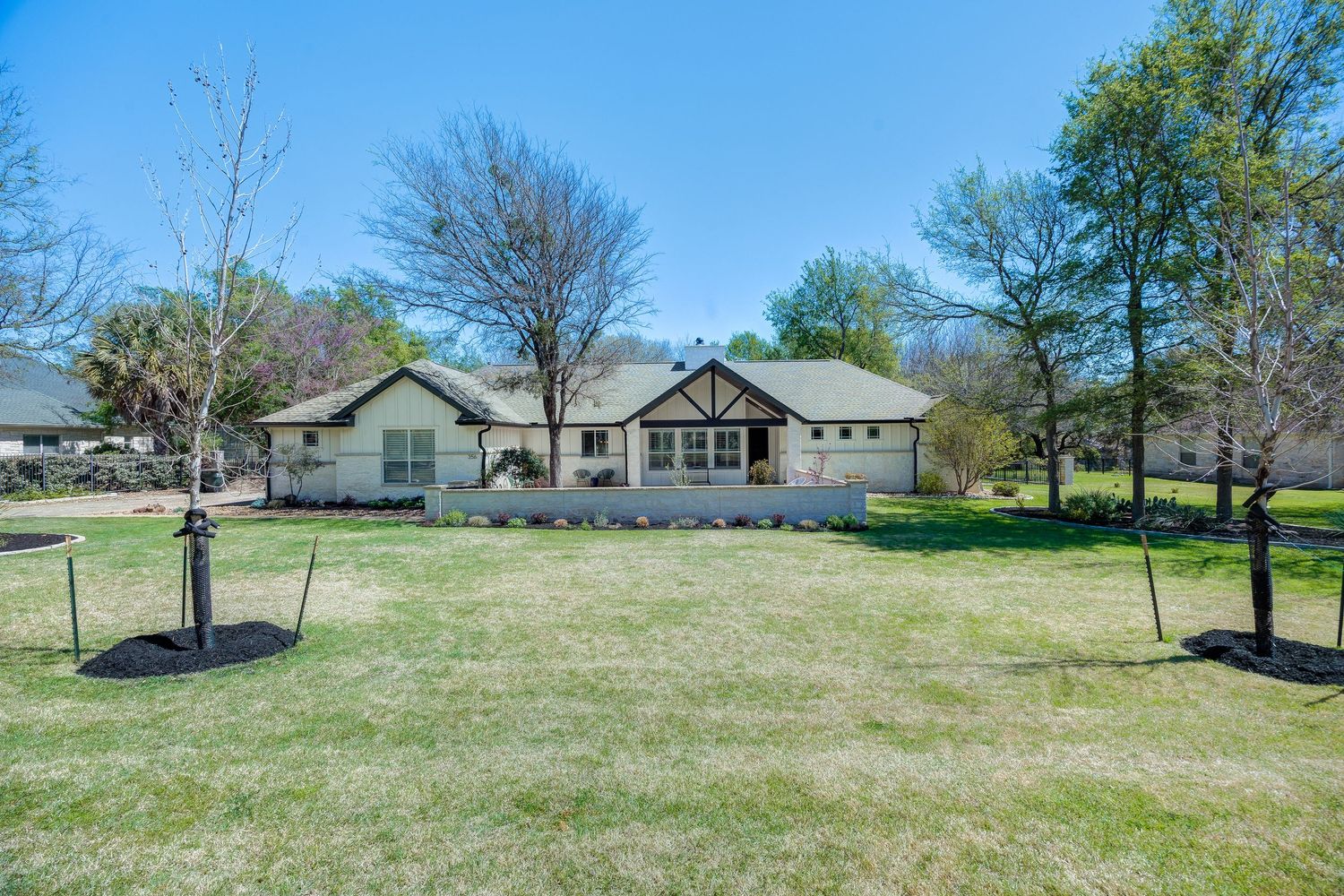 A charming ranch-style stone home with lush green lawn, mature trees, and manicured landscaping on a sunny spring day.