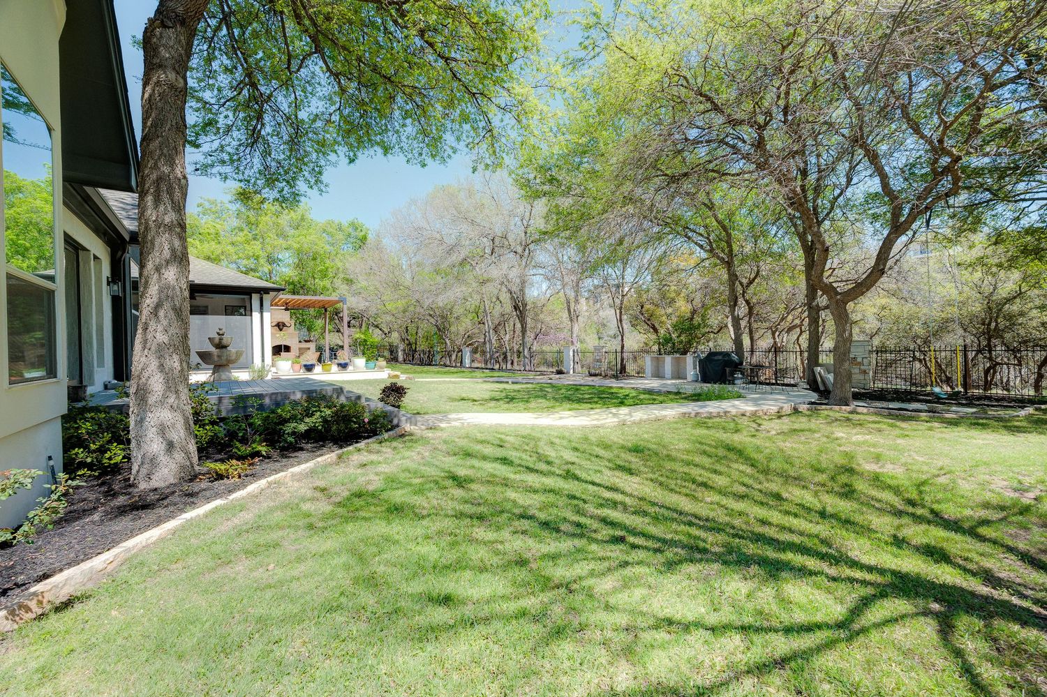 Lush green backyard with mature shade trees, manicured lawn, and a patio area surrounded by landscaped garden beds.