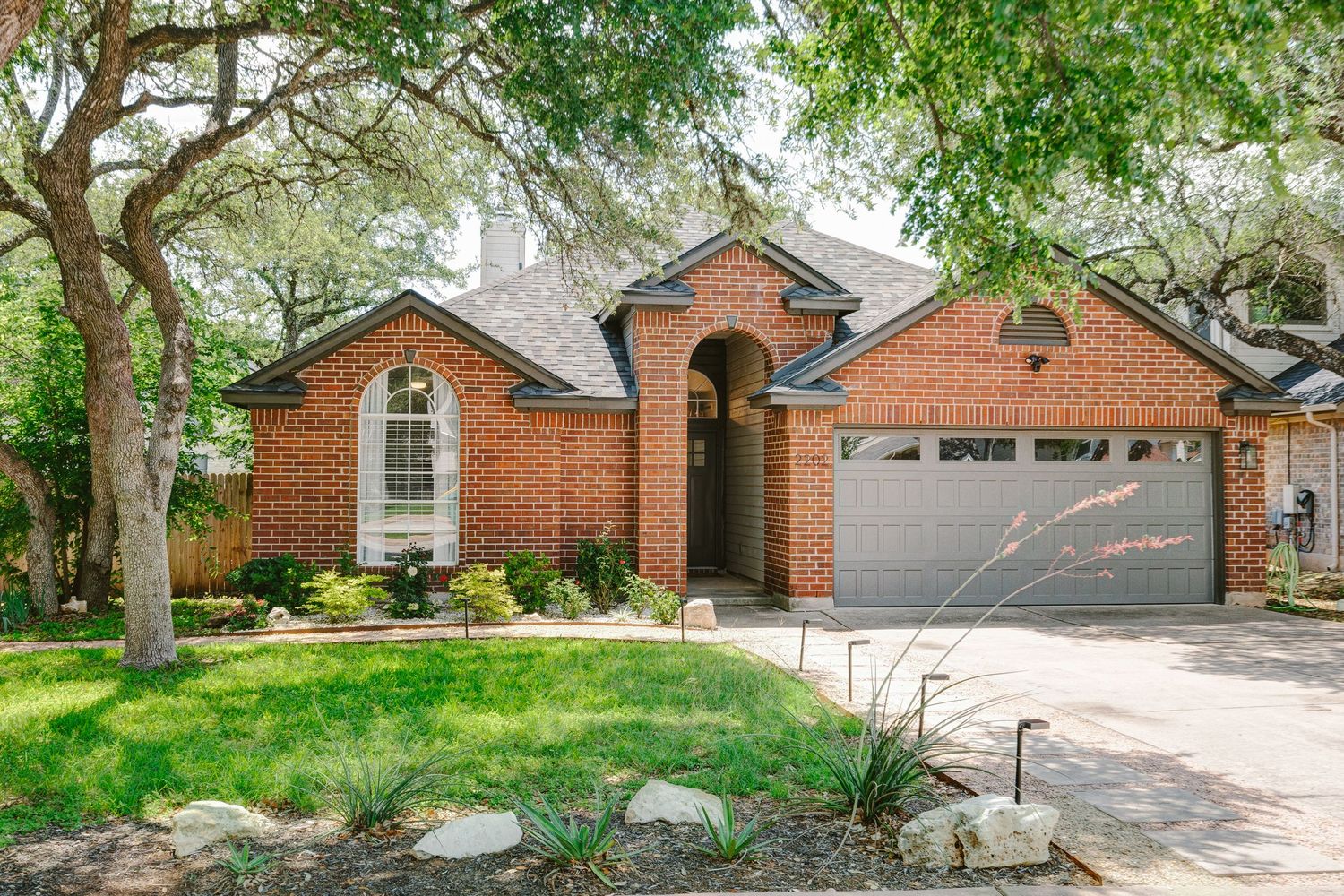 Brick ranch-style home with arched entryway, two-car garage, lush landscaping, and mature trees in a suburban neighborhood.