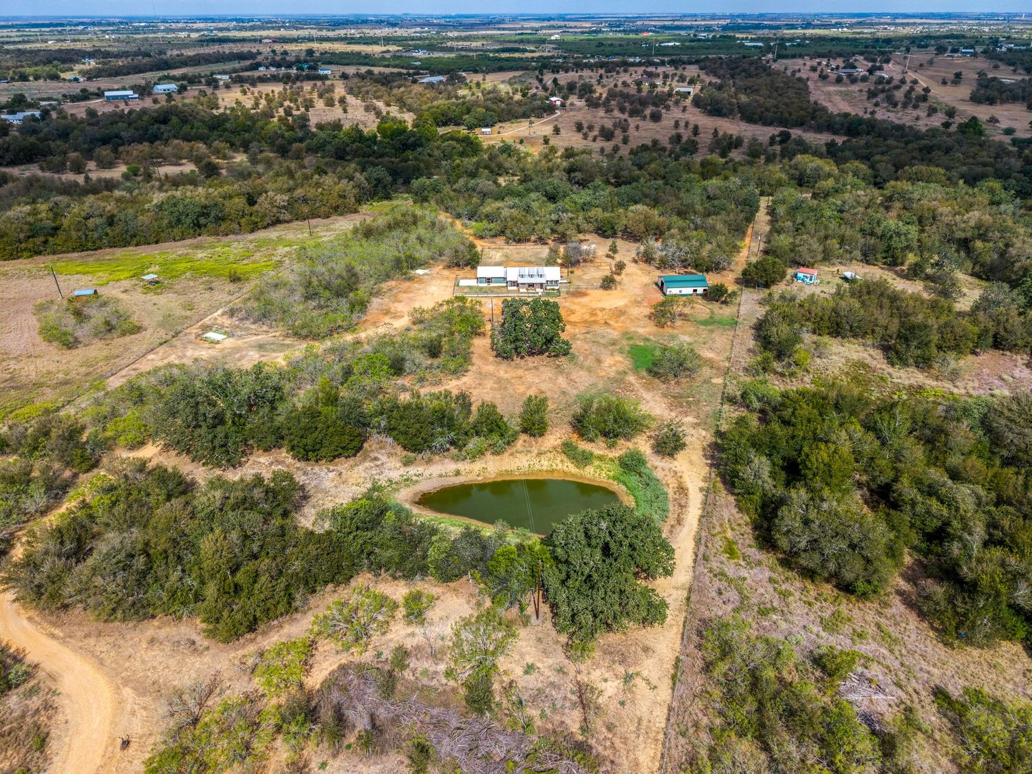Aerial view of a rural Texas property featuring a pond, scattered trees, dirt roads, and a white home surrounded by vast open land.