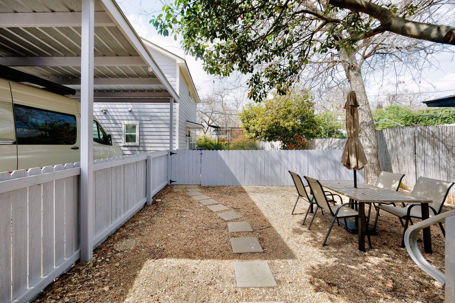 Backyard patio area with stepping stones, white fence, and outdoor dining furniture under a large tree.